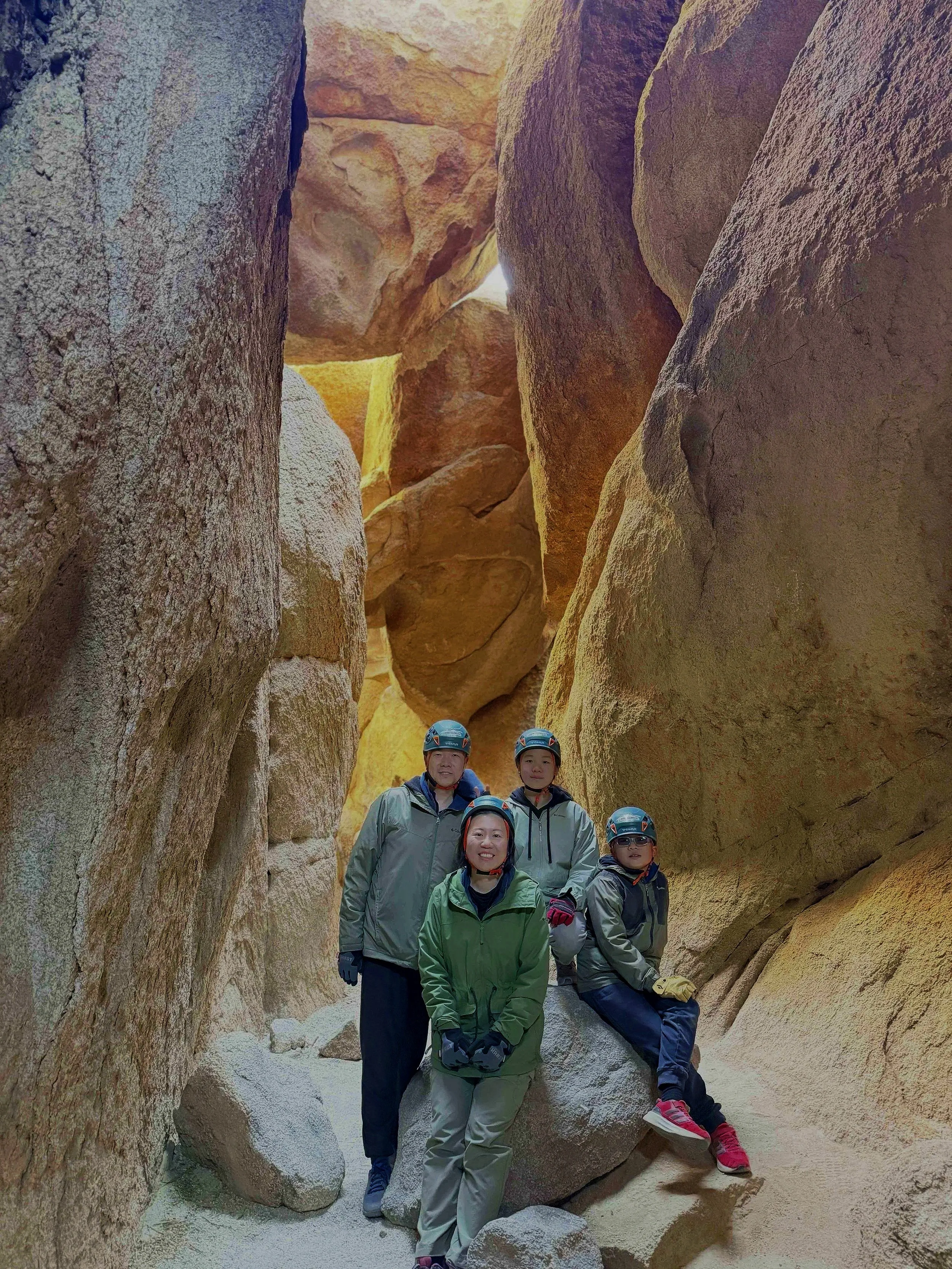 A group of five people wearing helmets and outdoor gear posing inside a narrow canyon with large, smooth rock walls with a small opening at the top allowing sunlight to illuminate the scene.