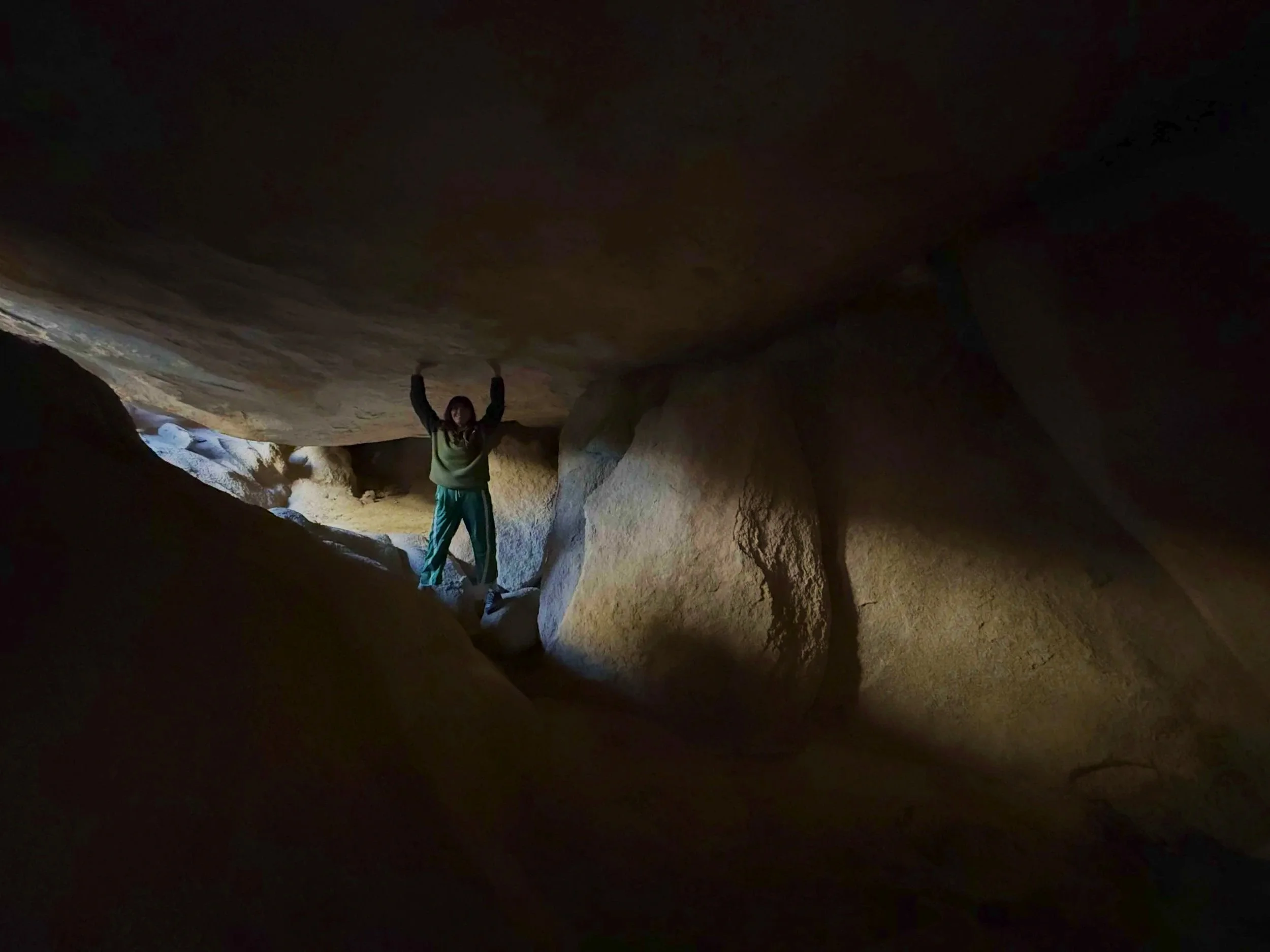 A person in a green jacket and blue pants standing inside a dark cave with large rock formations.