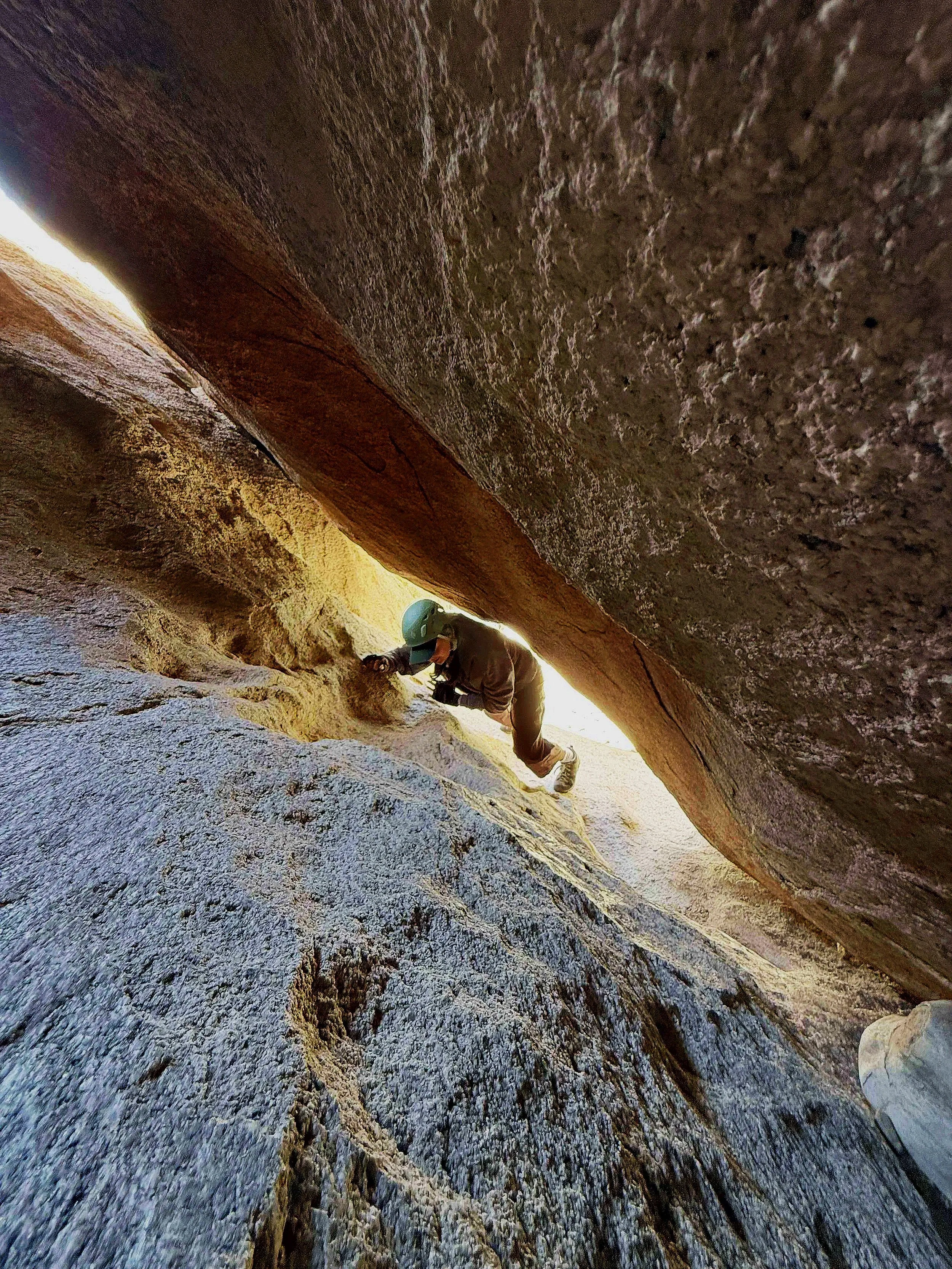 A person climbing through a narrow, rocky crevice in a mountain or canyon, wearing a helmet and outdoor clothing.