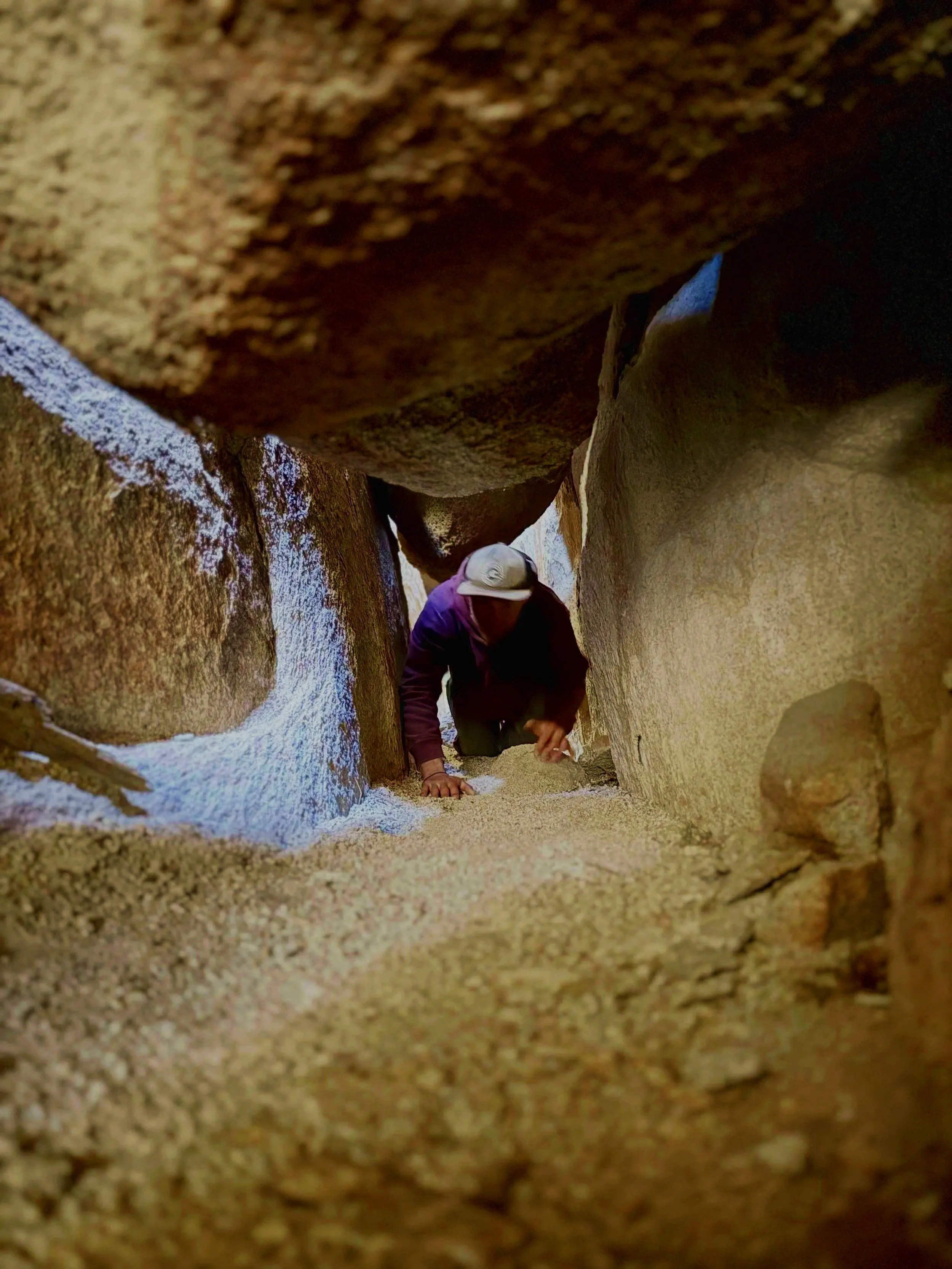 A person with a hat crawling through a narrow space between large rocks and sand.