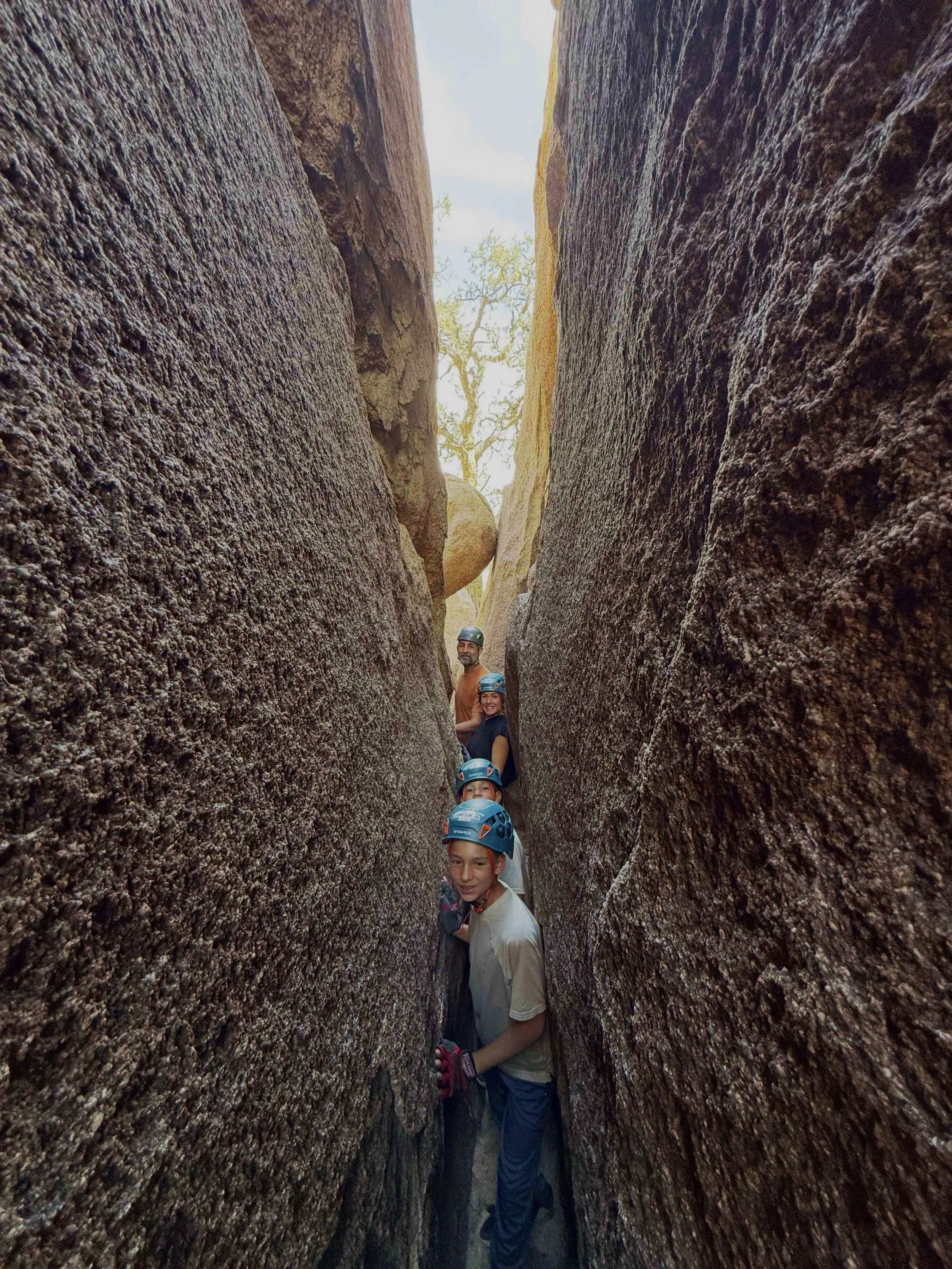 Group of children wearing helmets standing in a narrow crevice between two large rocks or boulders, with a tree in the background.