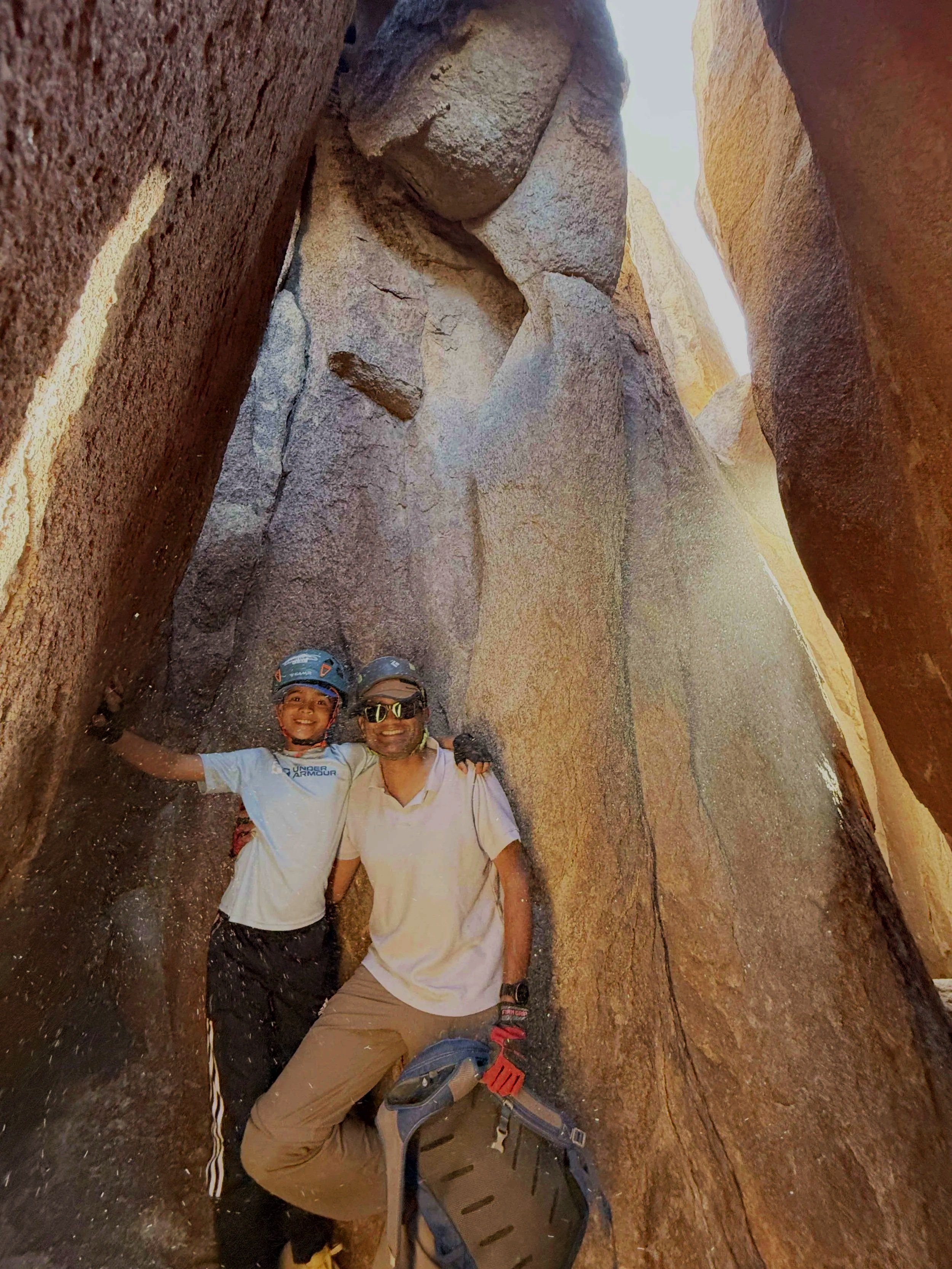 Two people, wearing helmets and casual outdoor clothing, standing inside a narrow, rocky canyon with large, textured rock walls on either side, smiling at the camera.