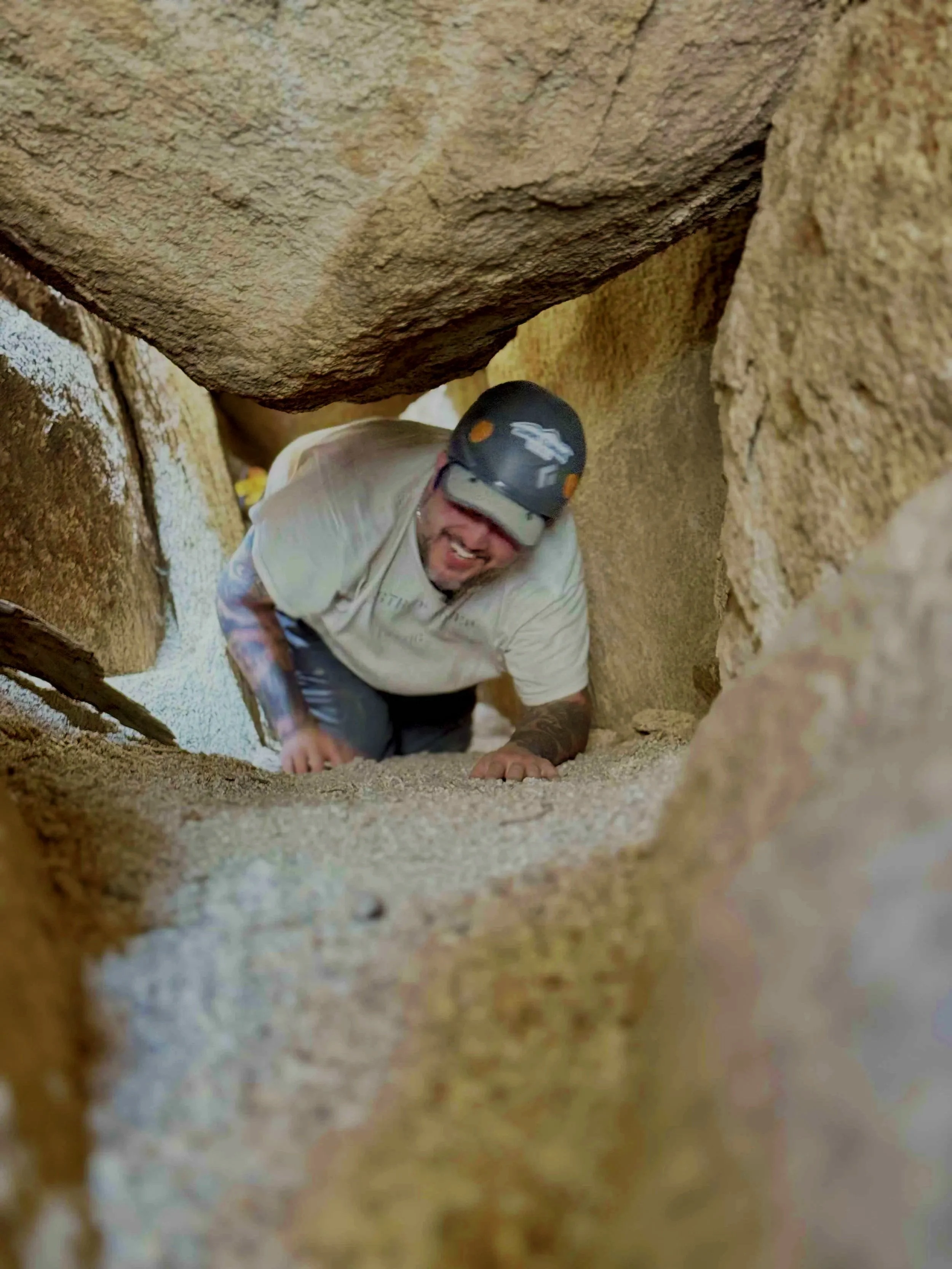 A person wearing a helmet crawling through a narrow crevice between large rocks in a cave.