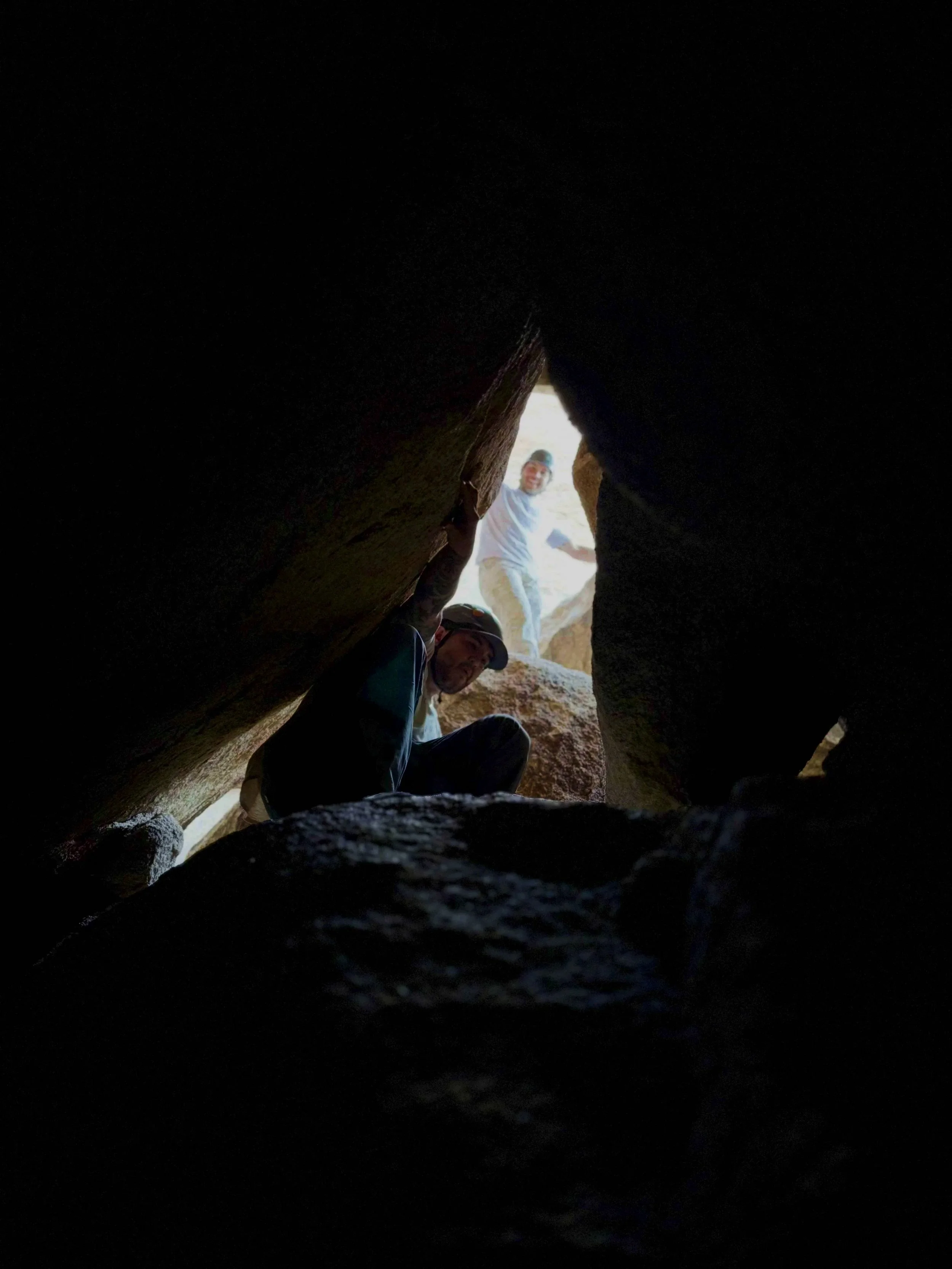 People exploring a narrow crevice or canyon with sunlight visible at the end, viewed from inside the gap.