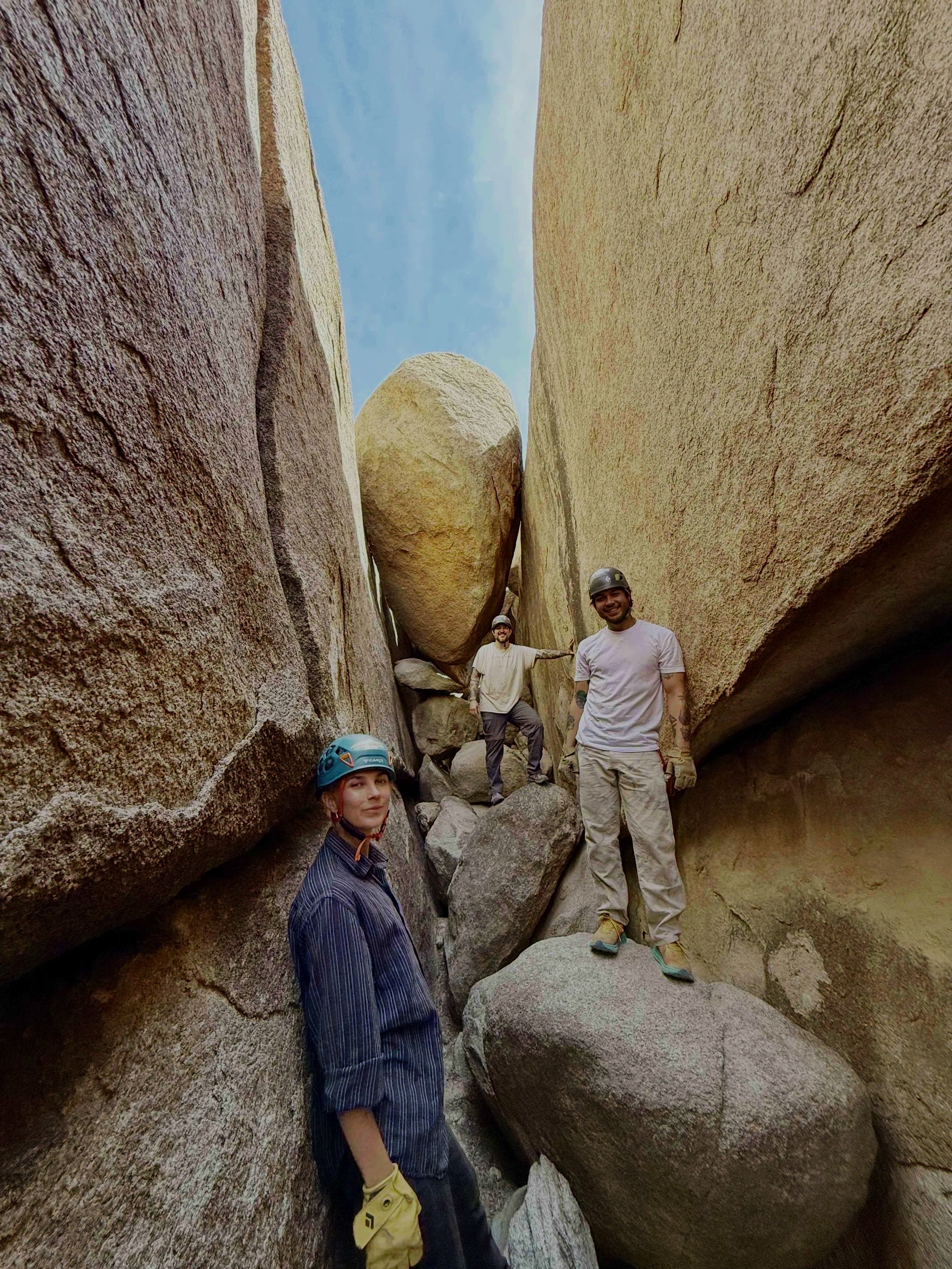 Three people standing between large rocks in a narrow canyon, some wearing helmets, one woman in the foreground with yellow gloves, two men in the background.