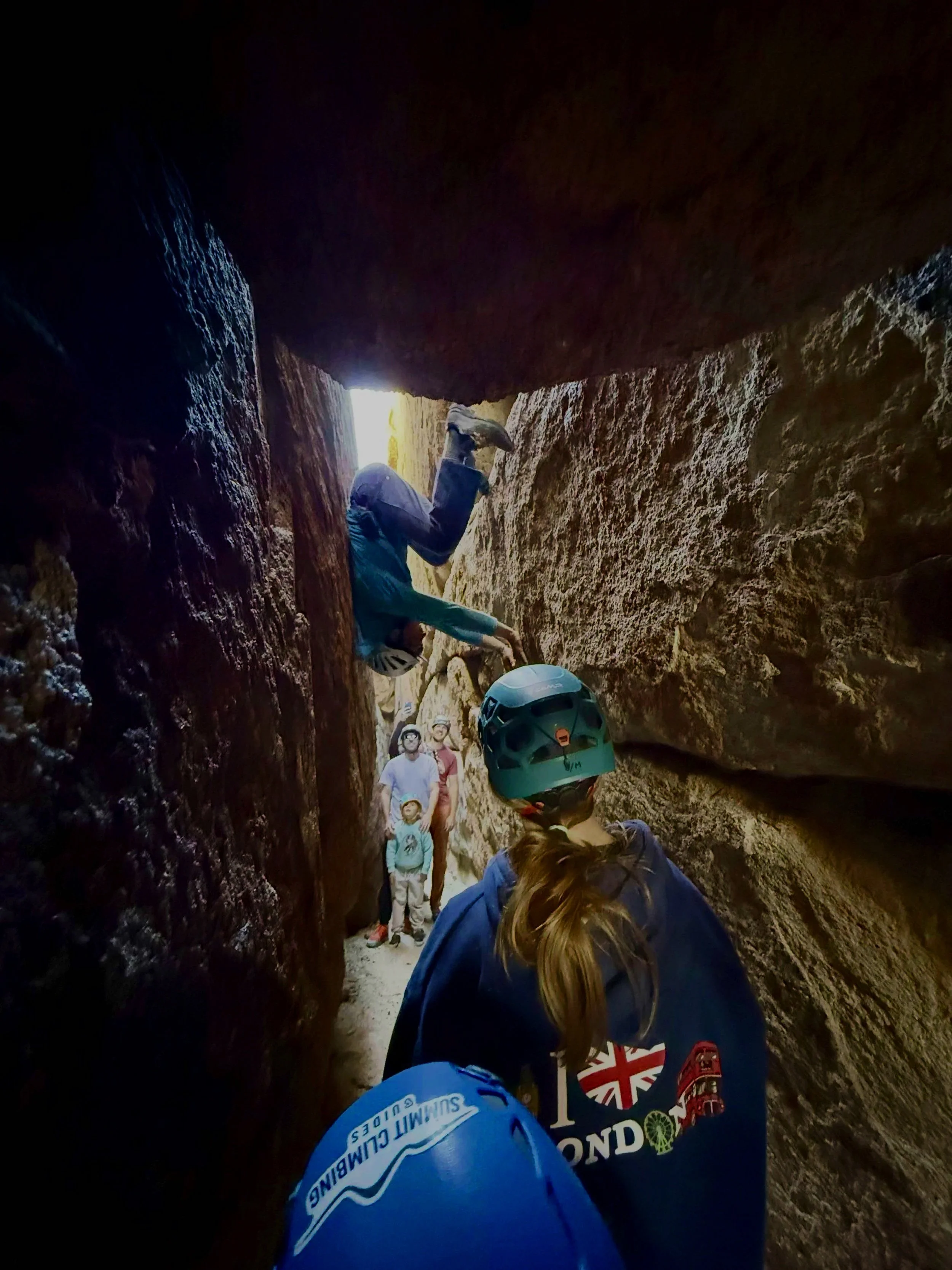 A group of people inside a narrow, rocky canyon with sunlight shining from behind the rocks. Some are wearing helmets and jackets, and appear to be involved in outdoor adventure or canyoning.