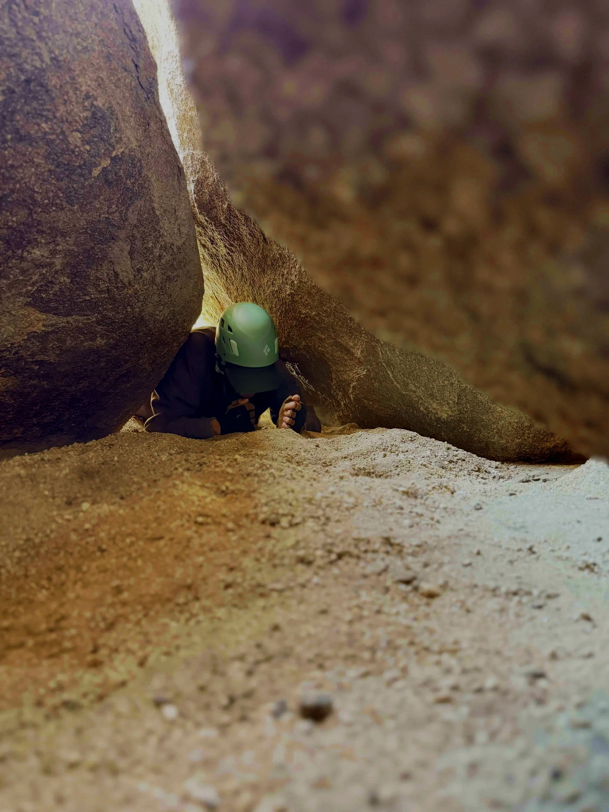 Climber in a helmet crawling through a narrow crevice between large rocks in a canyon or mountain area.