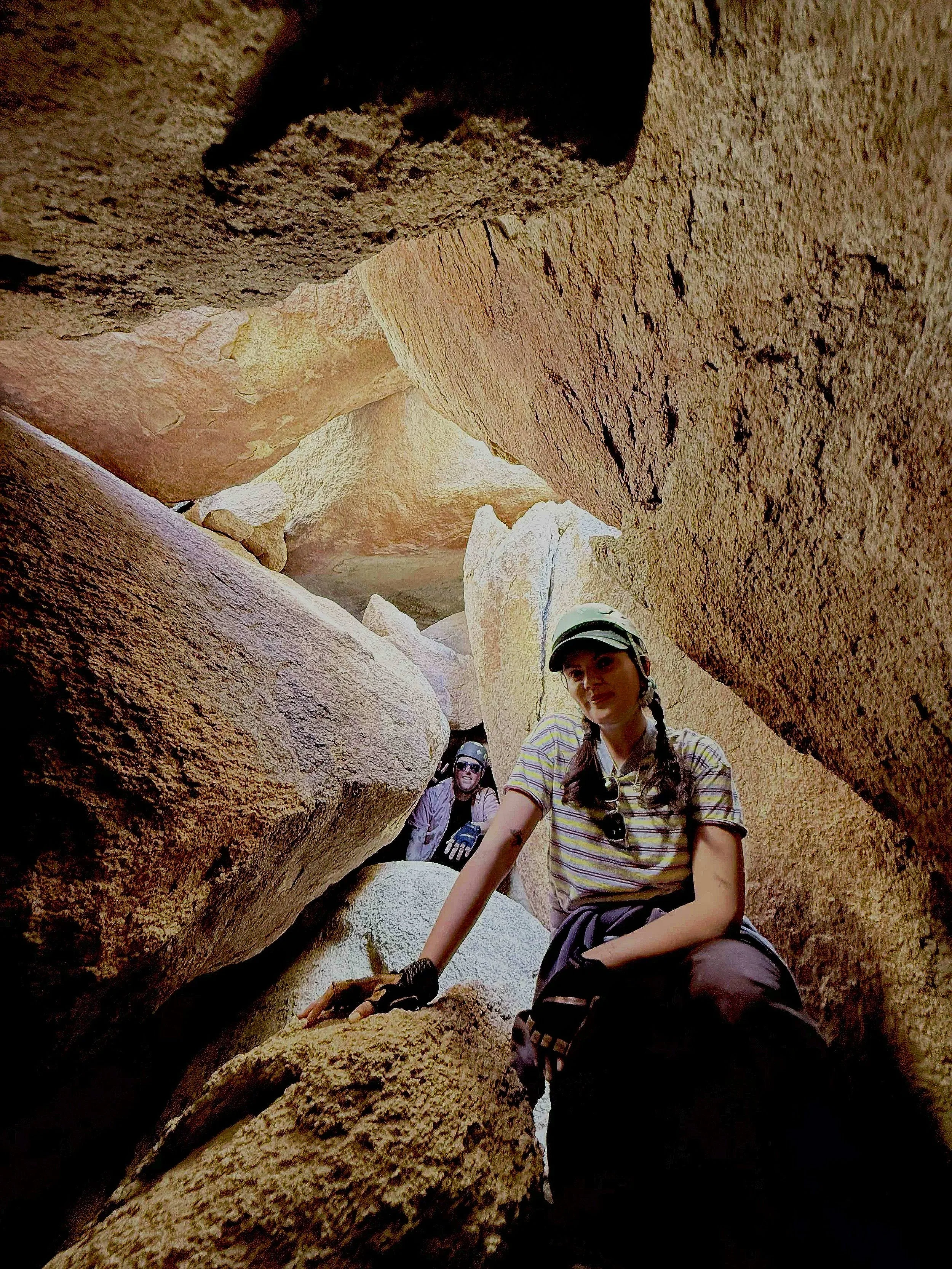 Two women inside a narrow, rocky canyon, climbing over large boulders, with one smiling at the camera. Both wear casual outdoor clothing and sunglasses, and one has a cap.
