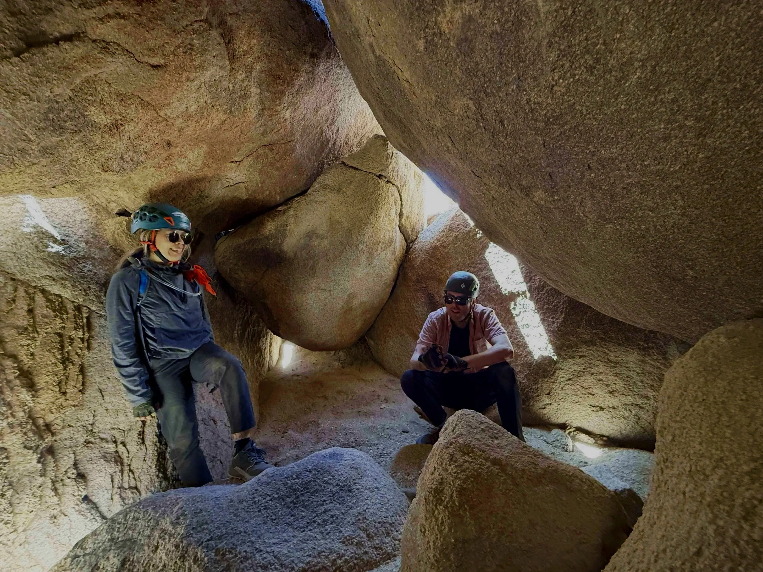 Three people wearing helmets and outdoor gear exploring inside a narrow, rocky canyon or crevice with large boulders and sunlight filtering through the top.