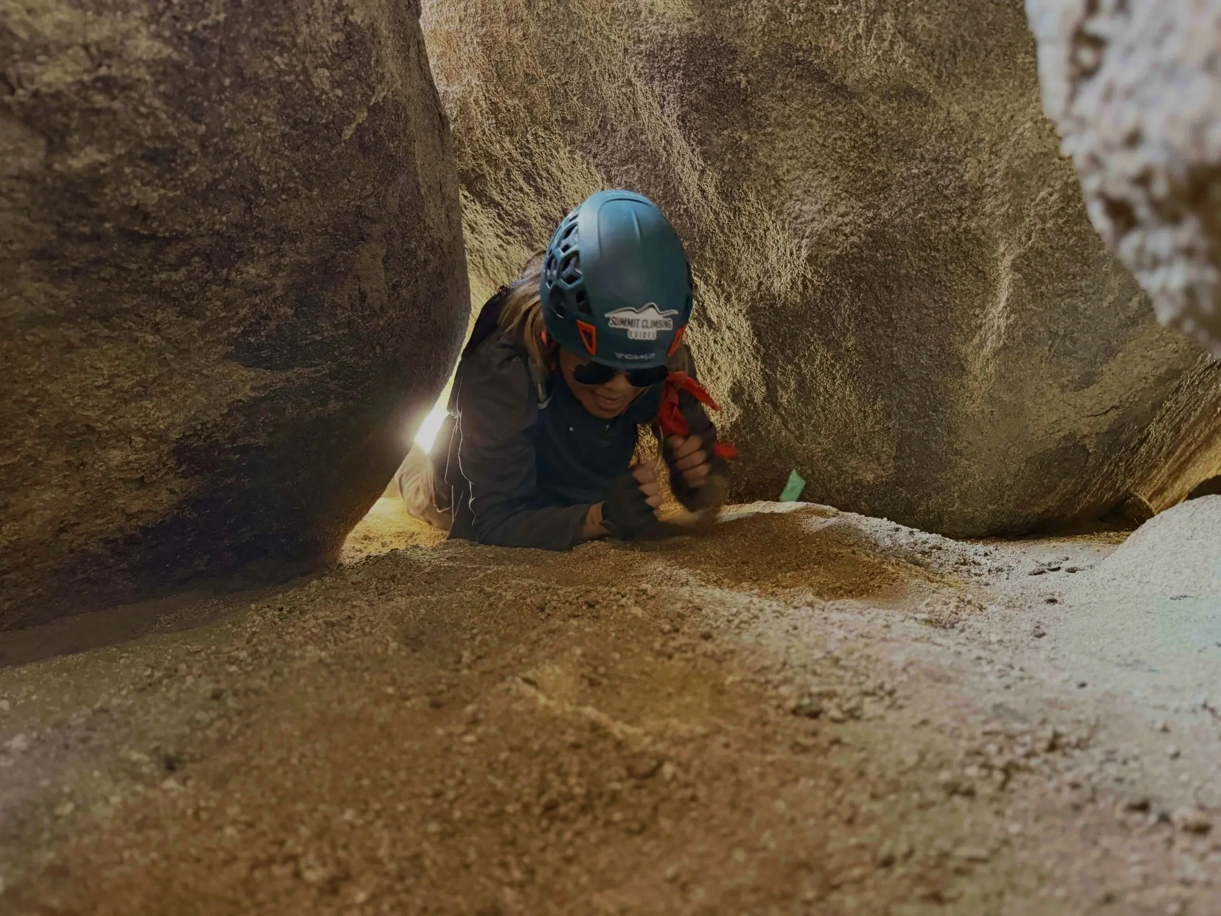 Person wearing a helmet and sunglasses crawling through a crevice in a rocky formation during an outdoor adventure.