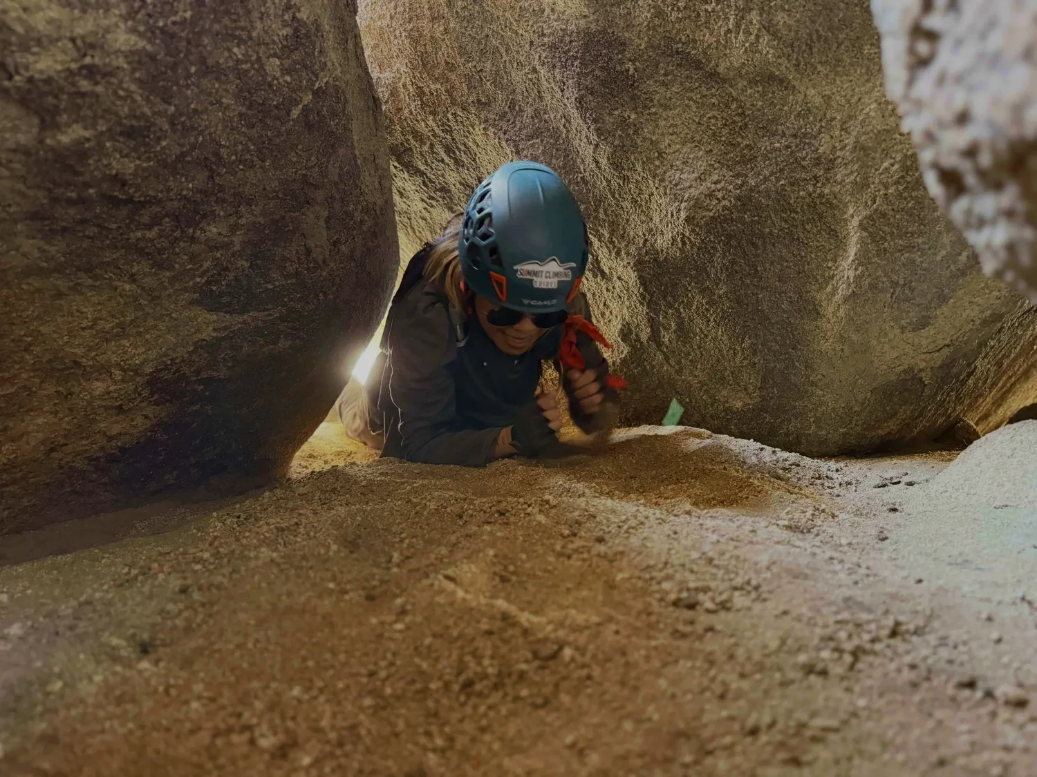 Person wearing a helmet and sunglasses crawling through a crevice in a rocky formation on The Mad Hatter exclusive route