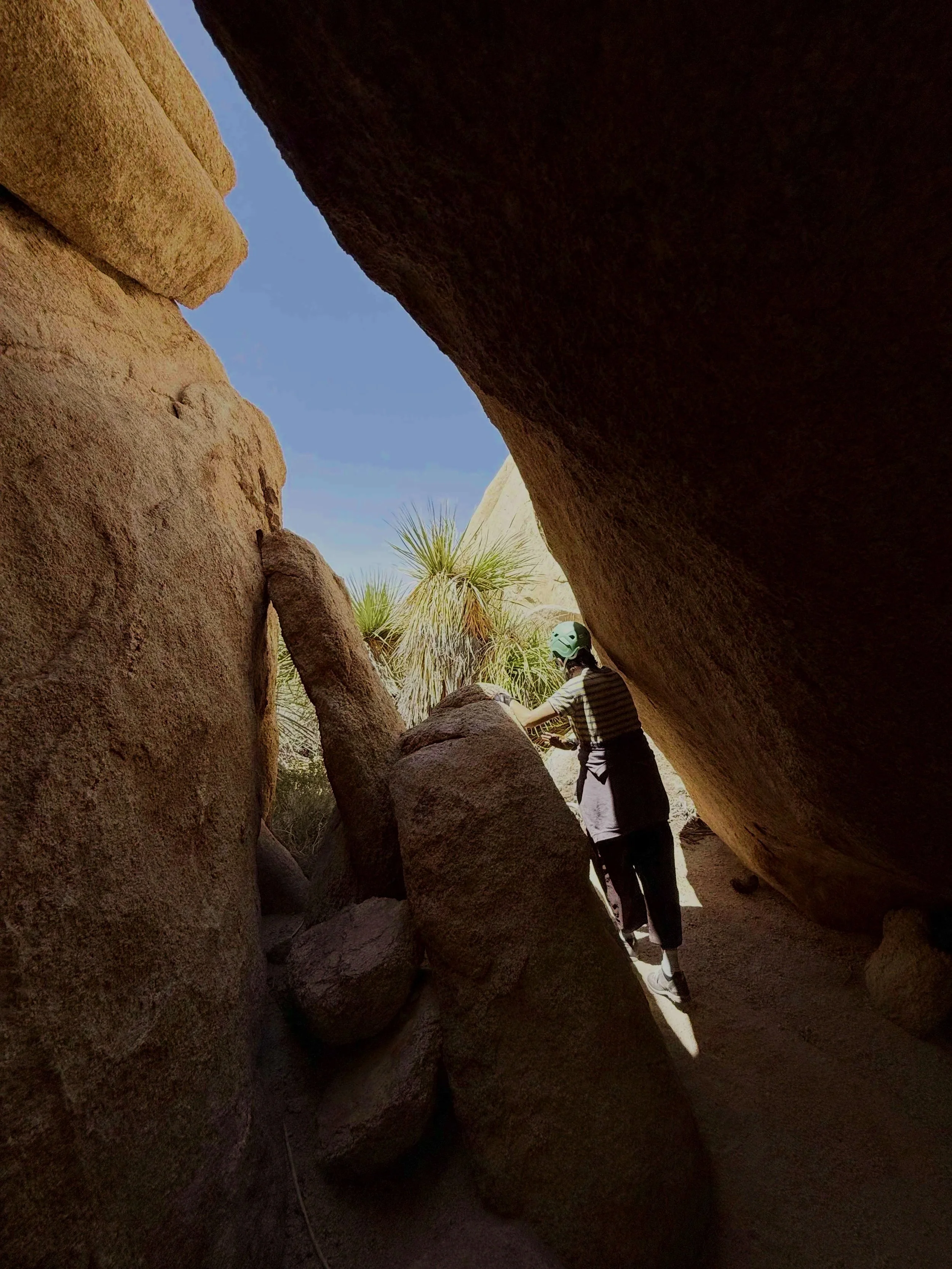 Person in striped shirt and helmet rock climbing in a narrow crevice between large boulders with desert plants in the background.