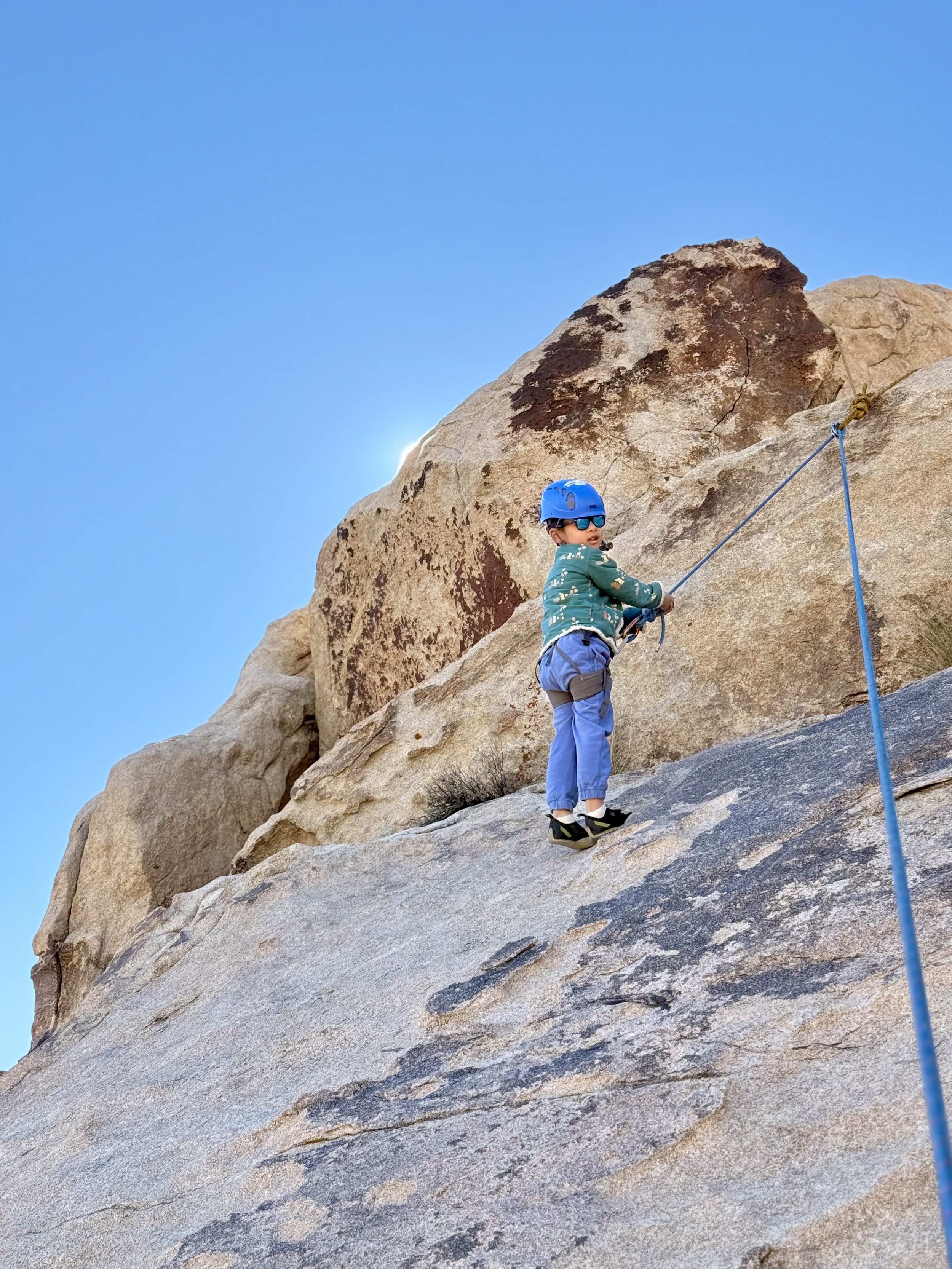 A young boy wearing a blue helmet and sunglasses, climbing and secured with a safety harness and rope on a rock face outdoors against a clear blue sky.
