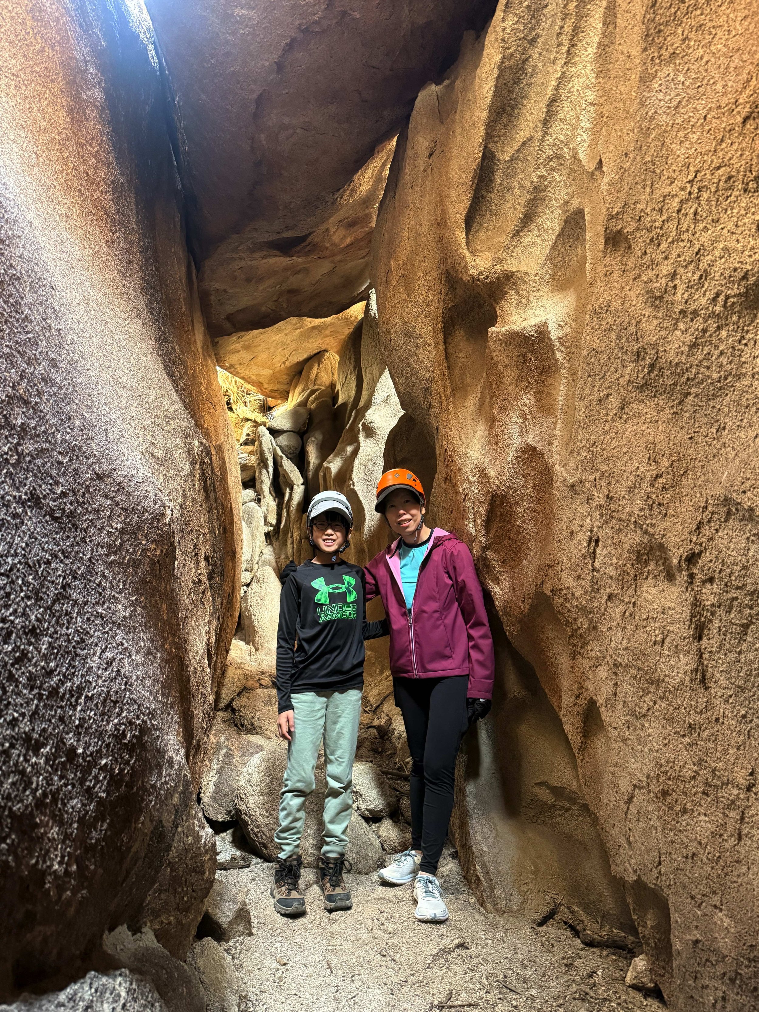 Two women wearing helmets and outdoor clothing standing inside narrow rocky canyon with large beige and reddish rocks surrounding them.