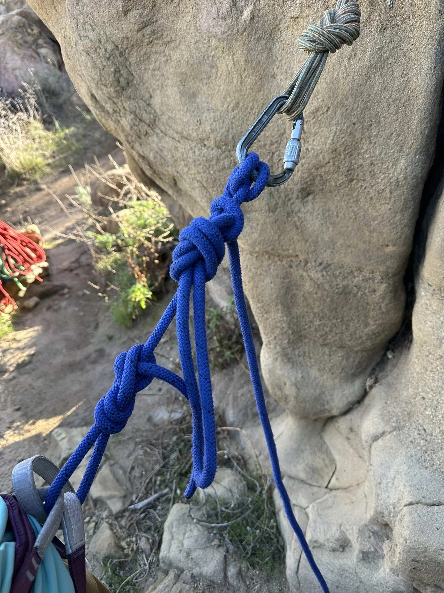 Climbing gear attached to a rock face, including a carabiner and a blue knot tied with a blue rope, with a backpack at the bottom left.