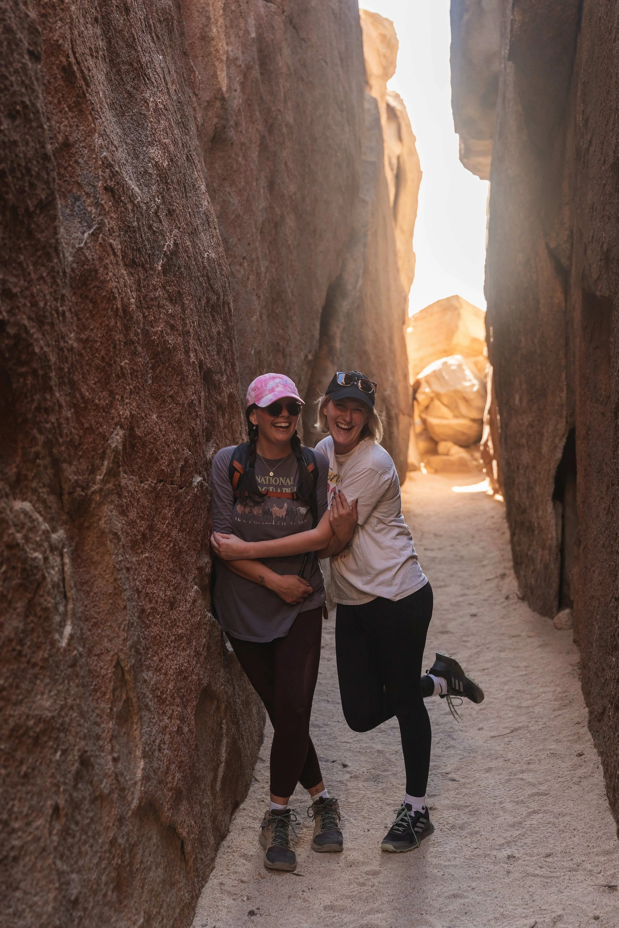 Two women smiling and hugging in a narrow rocky canyon with large boulders and sunlight at the end of the valley.