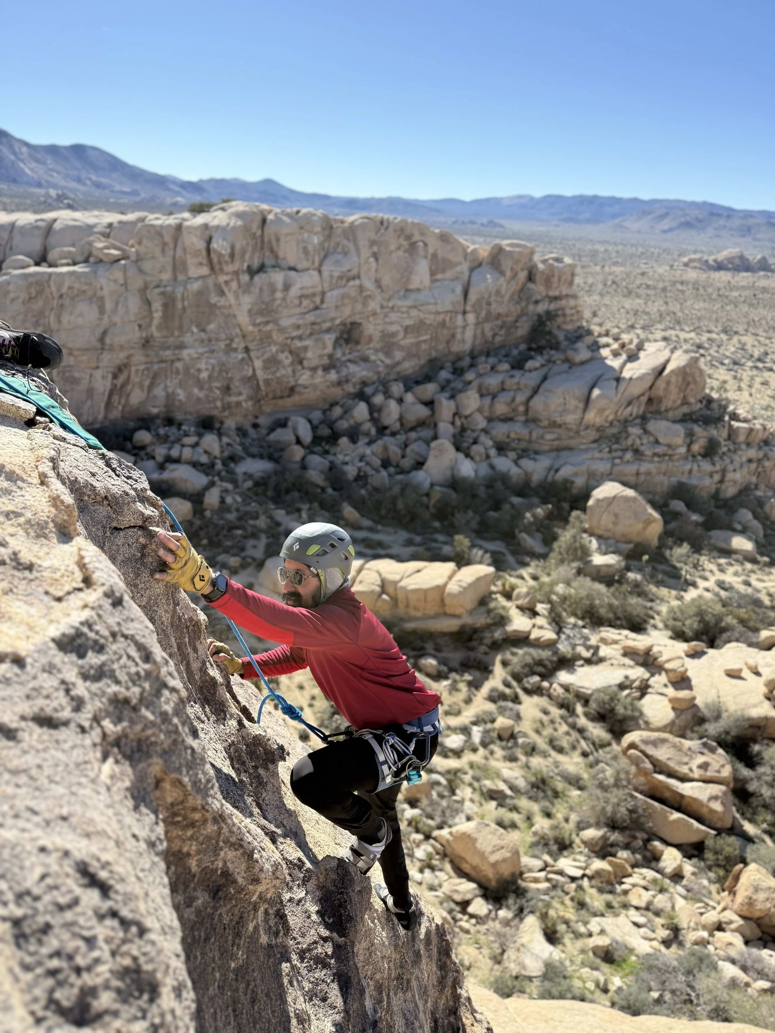 A man rock climbing on a steep desert rock face with desert landscape and mountains in the background.