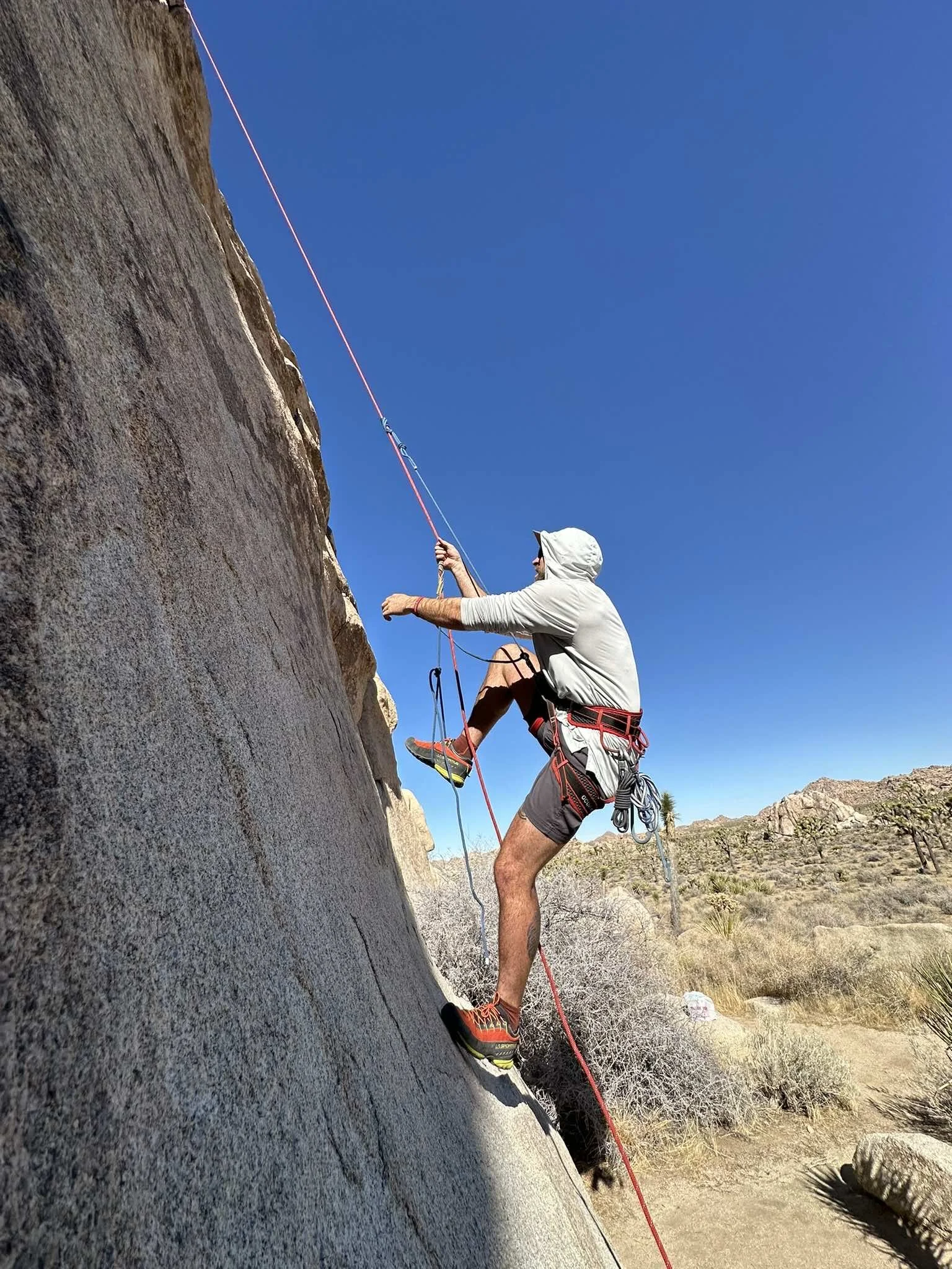 A person in a gray hoodie, black shorts, and climbing shoes is rock climbing on a steep outdoor granite wall under a clear blue sky, using safety gear and ropes.