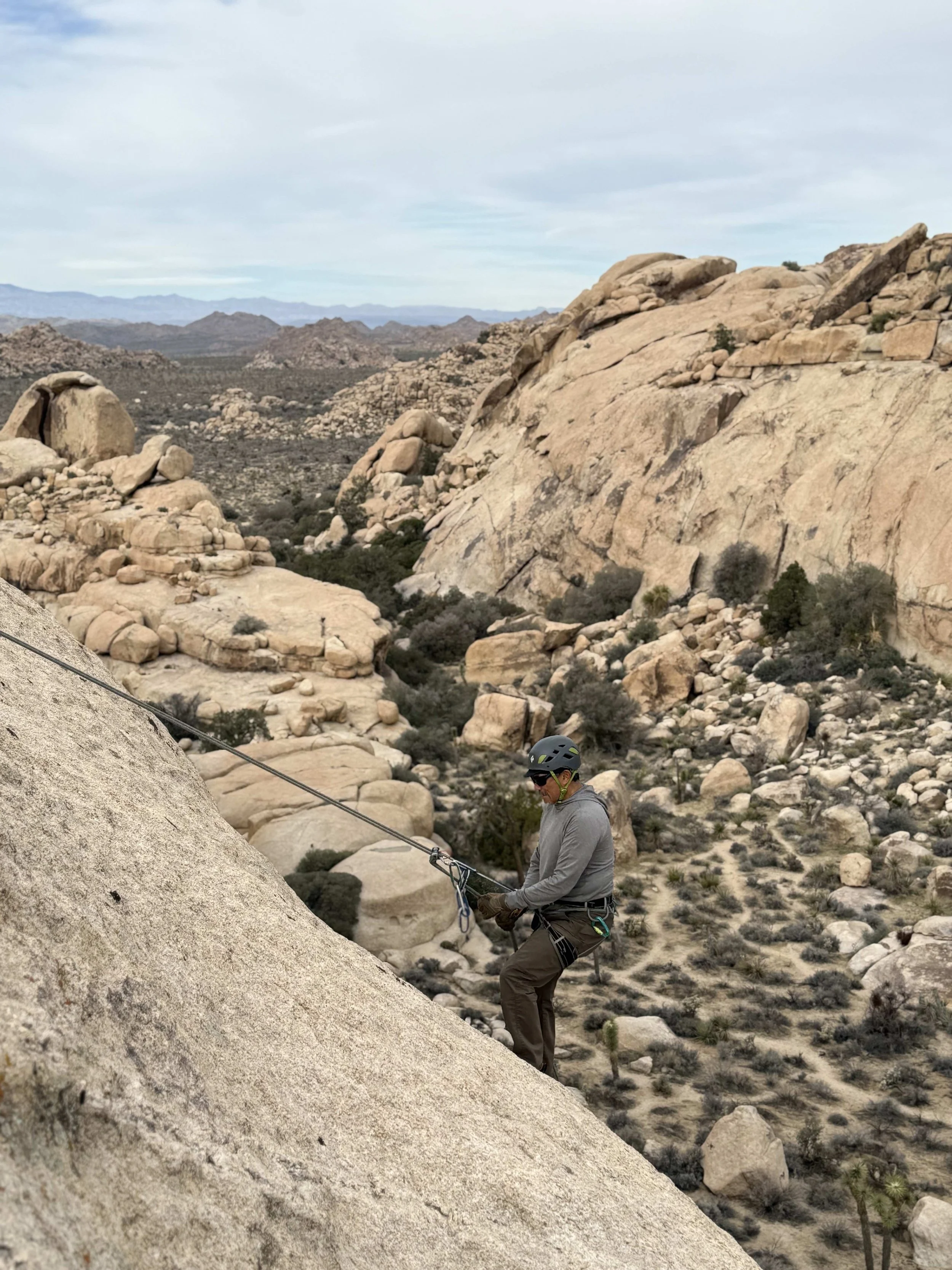 A person rock climbing on a steep granite slope in a desert landscape with large boulders and rocky hills in the background.