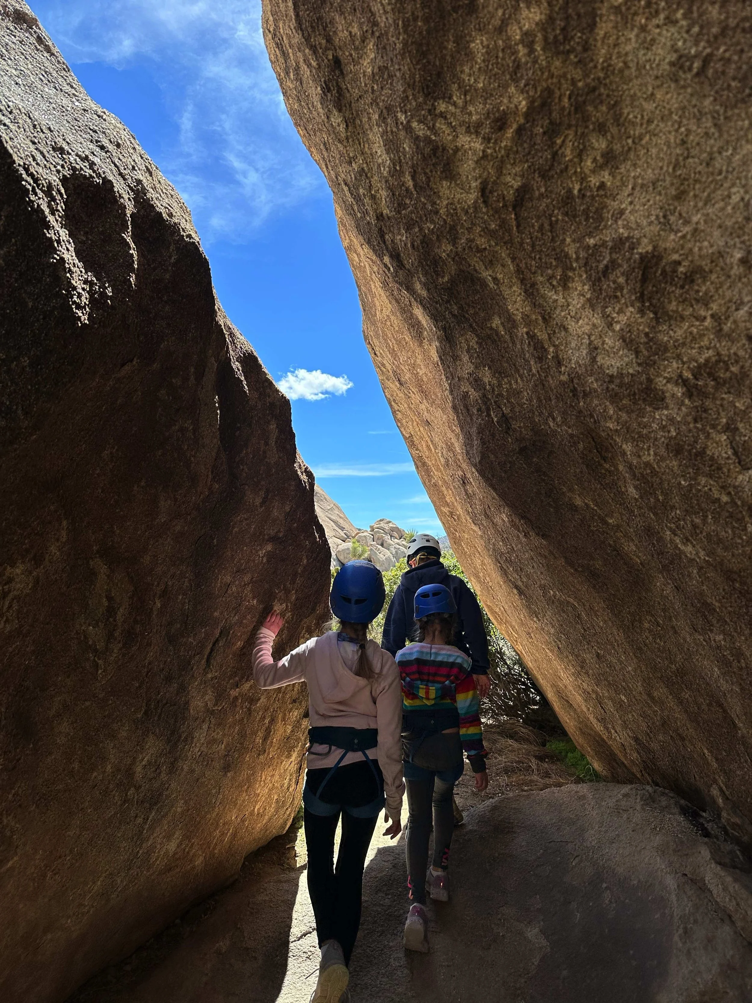 Group of children and an adult exploring a rock formation with large boulders, wearing helmets outdoors on a sunny day.