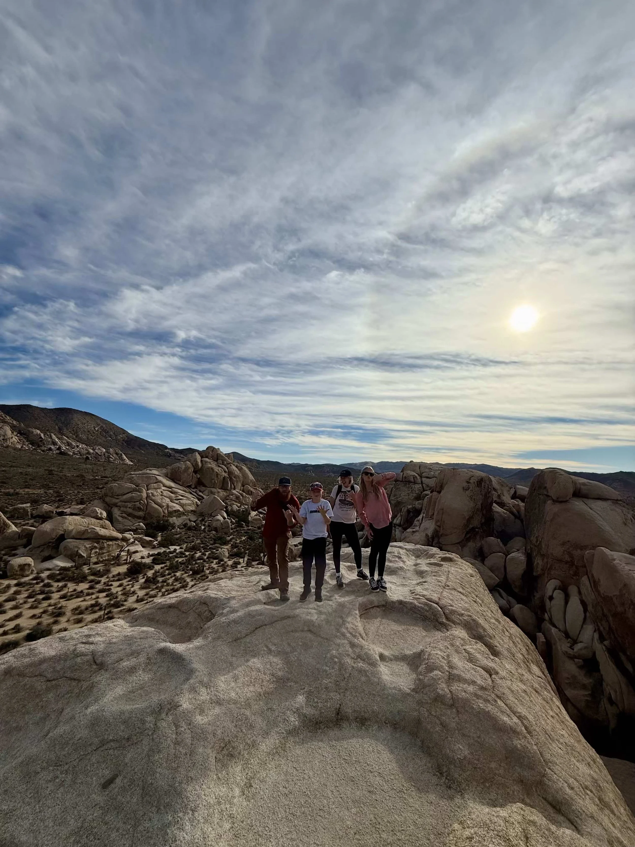 Four people standing on a large rock formation in a desert landscape with rocky hills, sparse vegetation, and a partly cloudy sky with the sun shining.