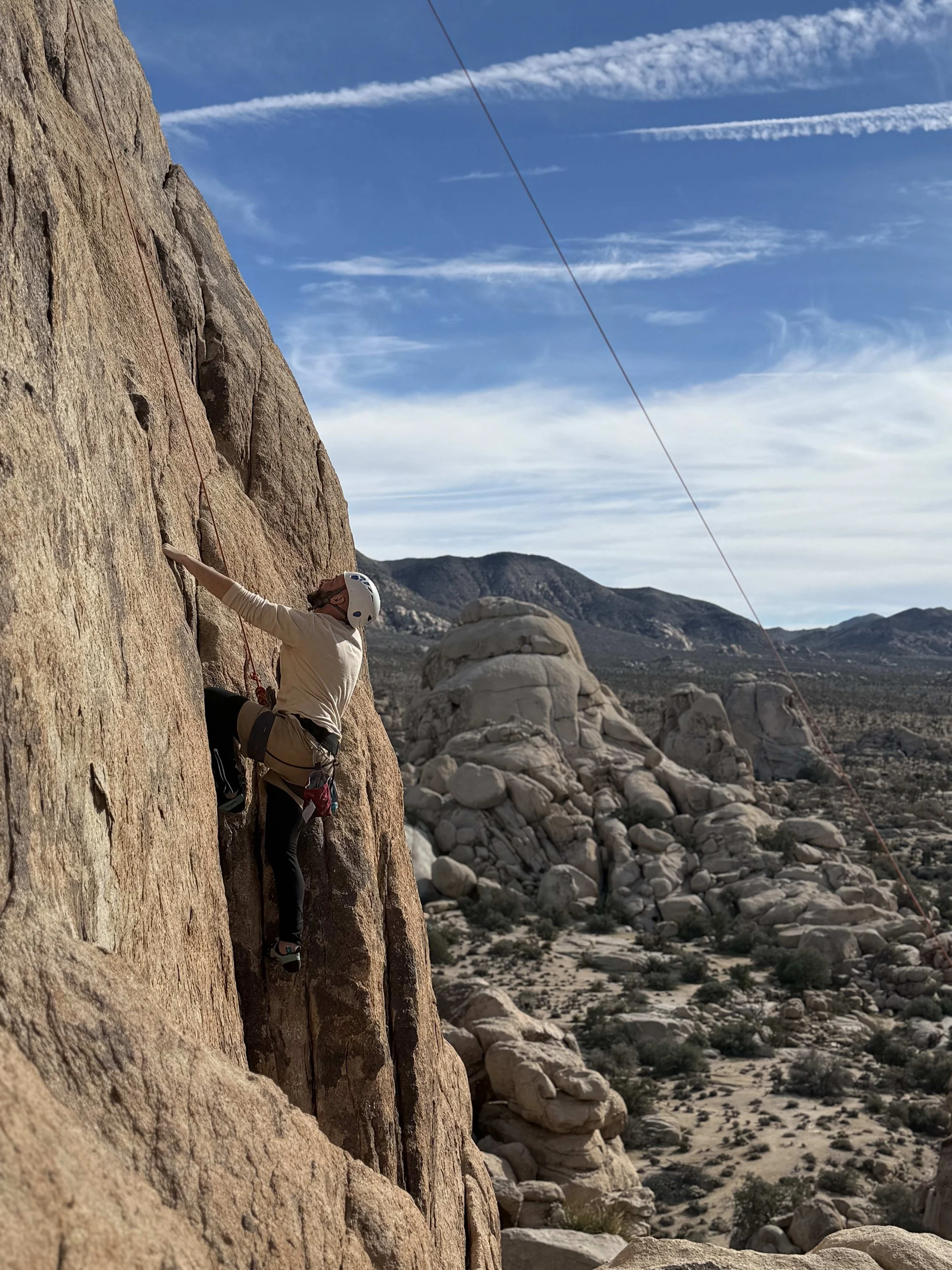 A person rock climbing on a steep granite cliff with desert landscape and mountains in the background under a partly cloudy sky.