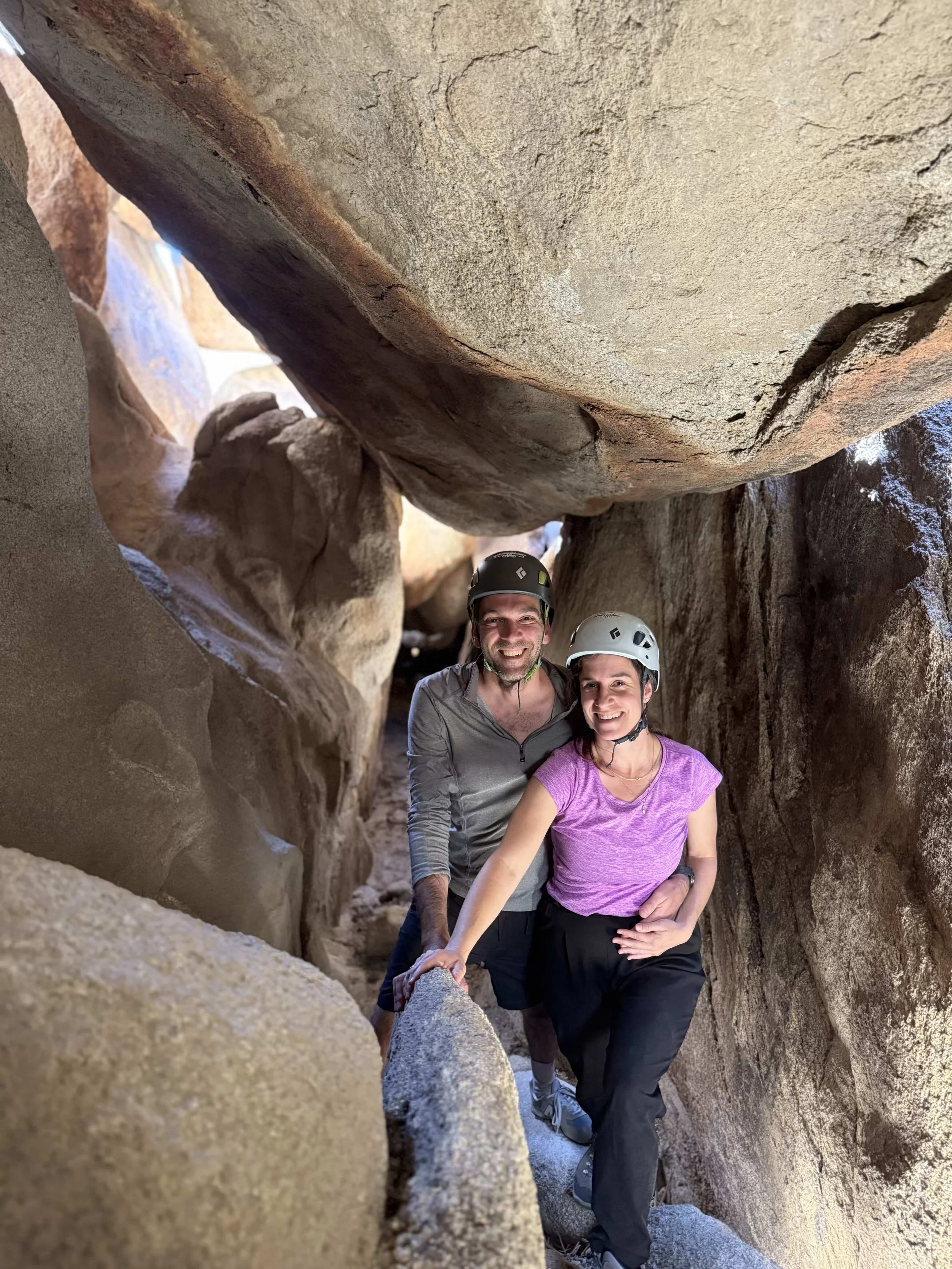 Two hikers wearing helmets and outdoor clothing exploring a narrow rock crevice, surrounded by large rocks and boulders.