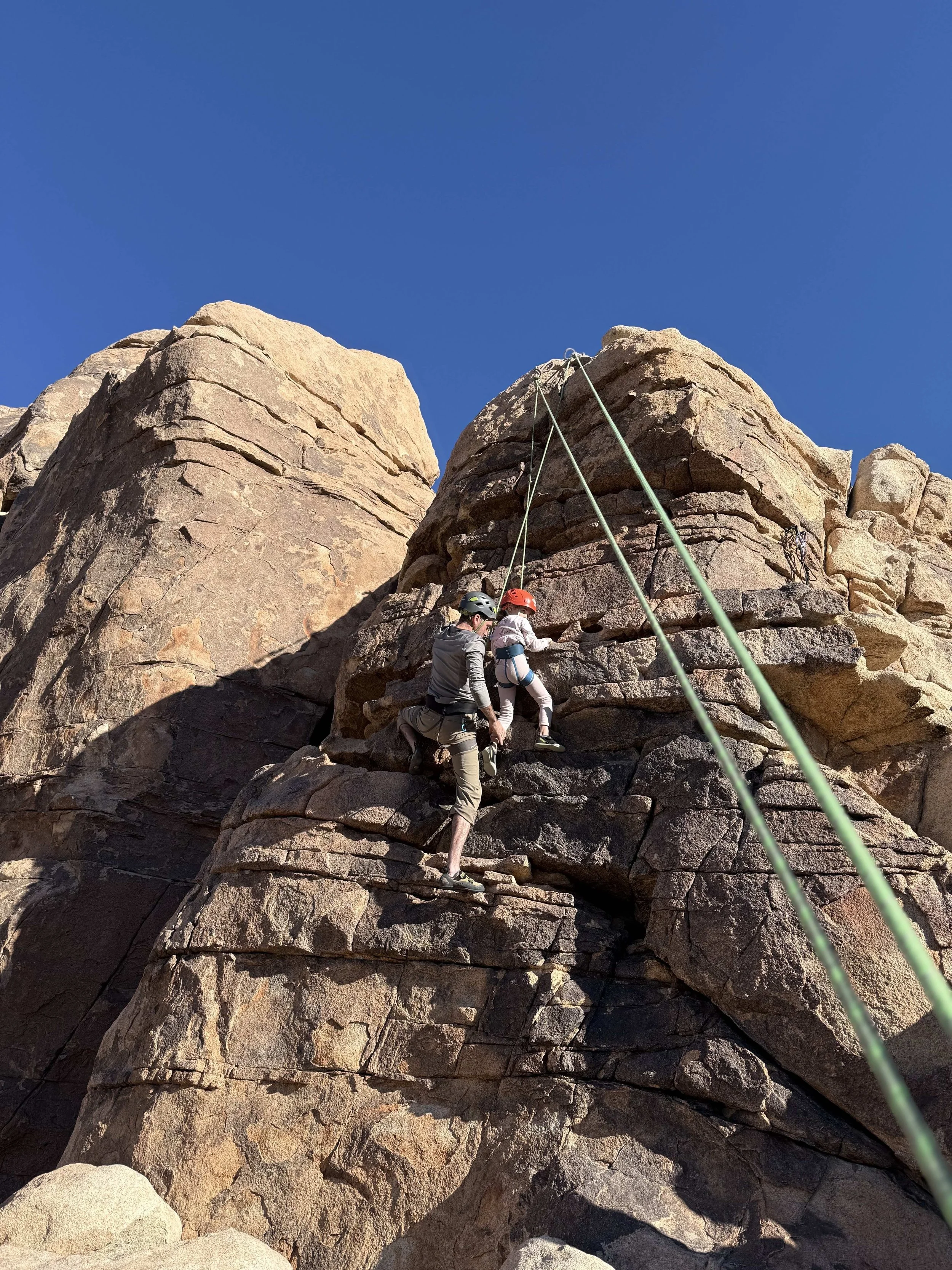 Two people rock climbing on a large rocky cliff with safety ropes and helmets against a clear blue sky.