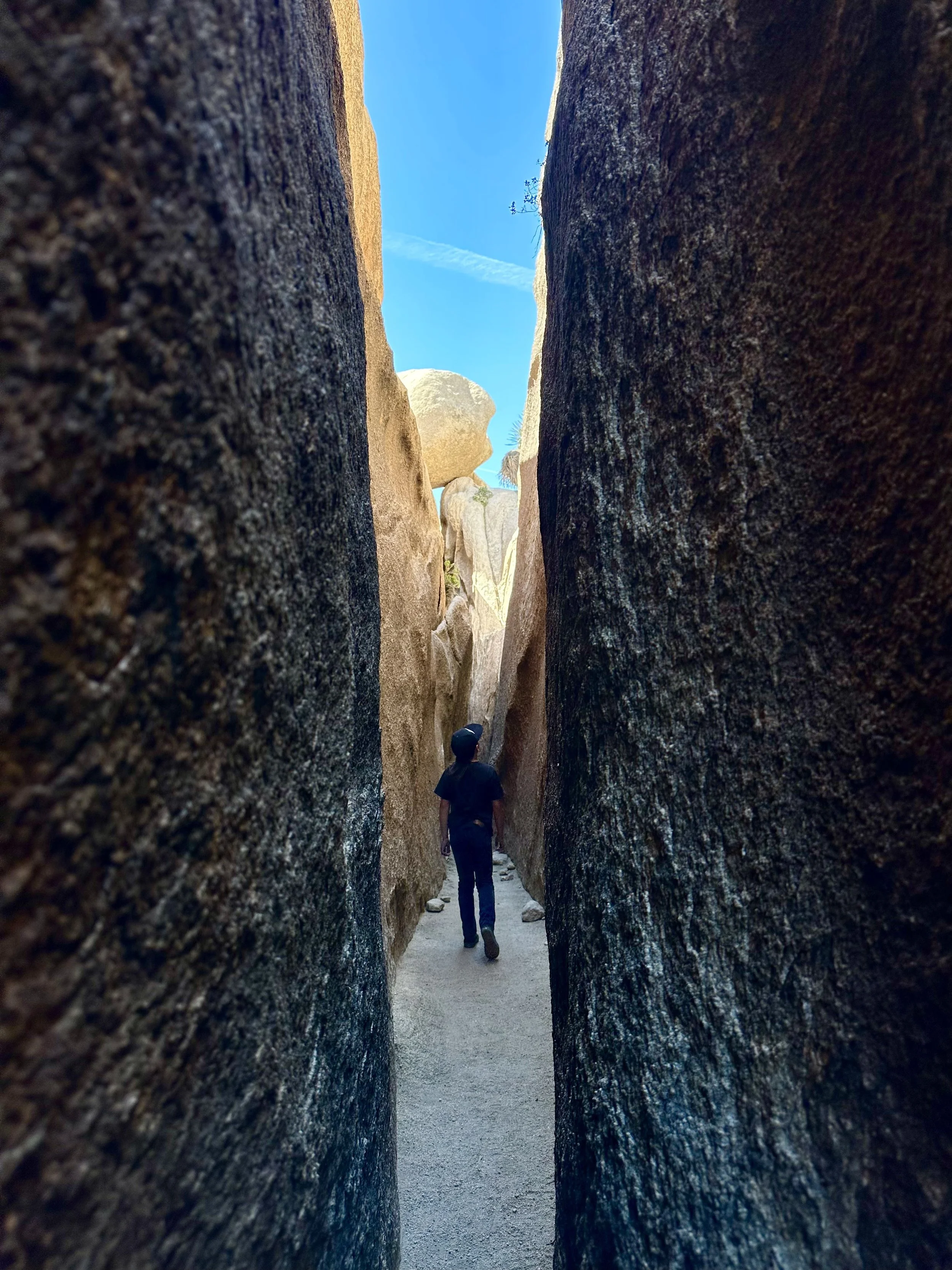 Person hiking between tall rock formations on a narrow trail, with a blue sky overhead.