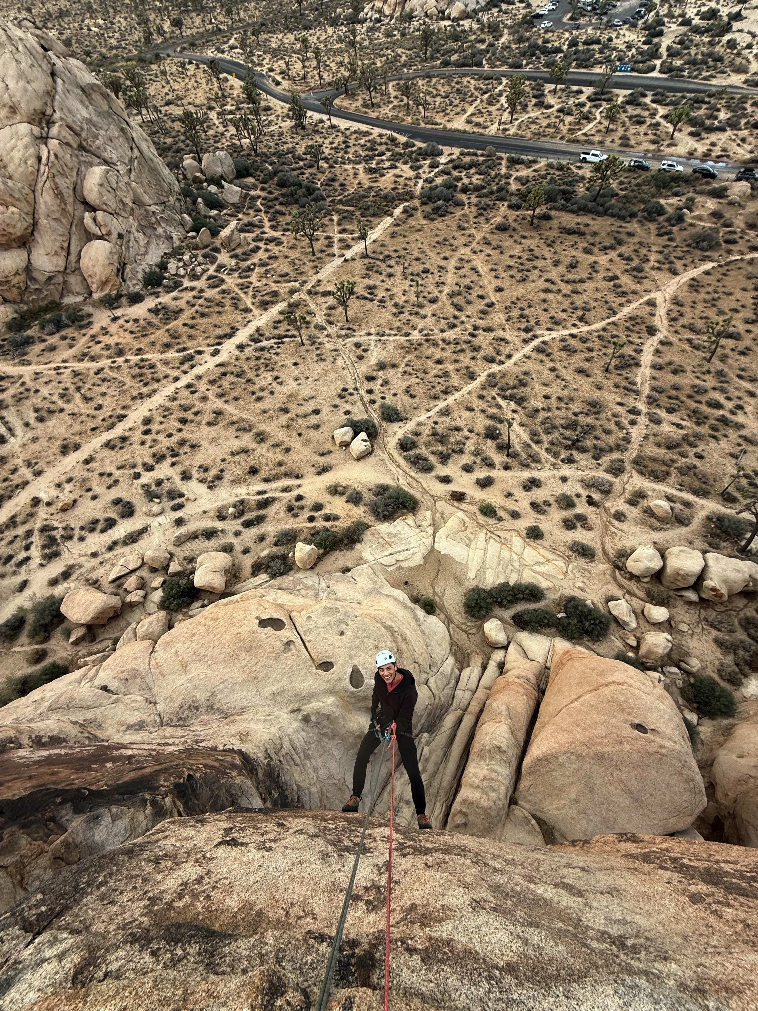 Person wearing a helmet climbing a rock formation with a view of desert terrain, scattered bushes, and a winding road in the background.