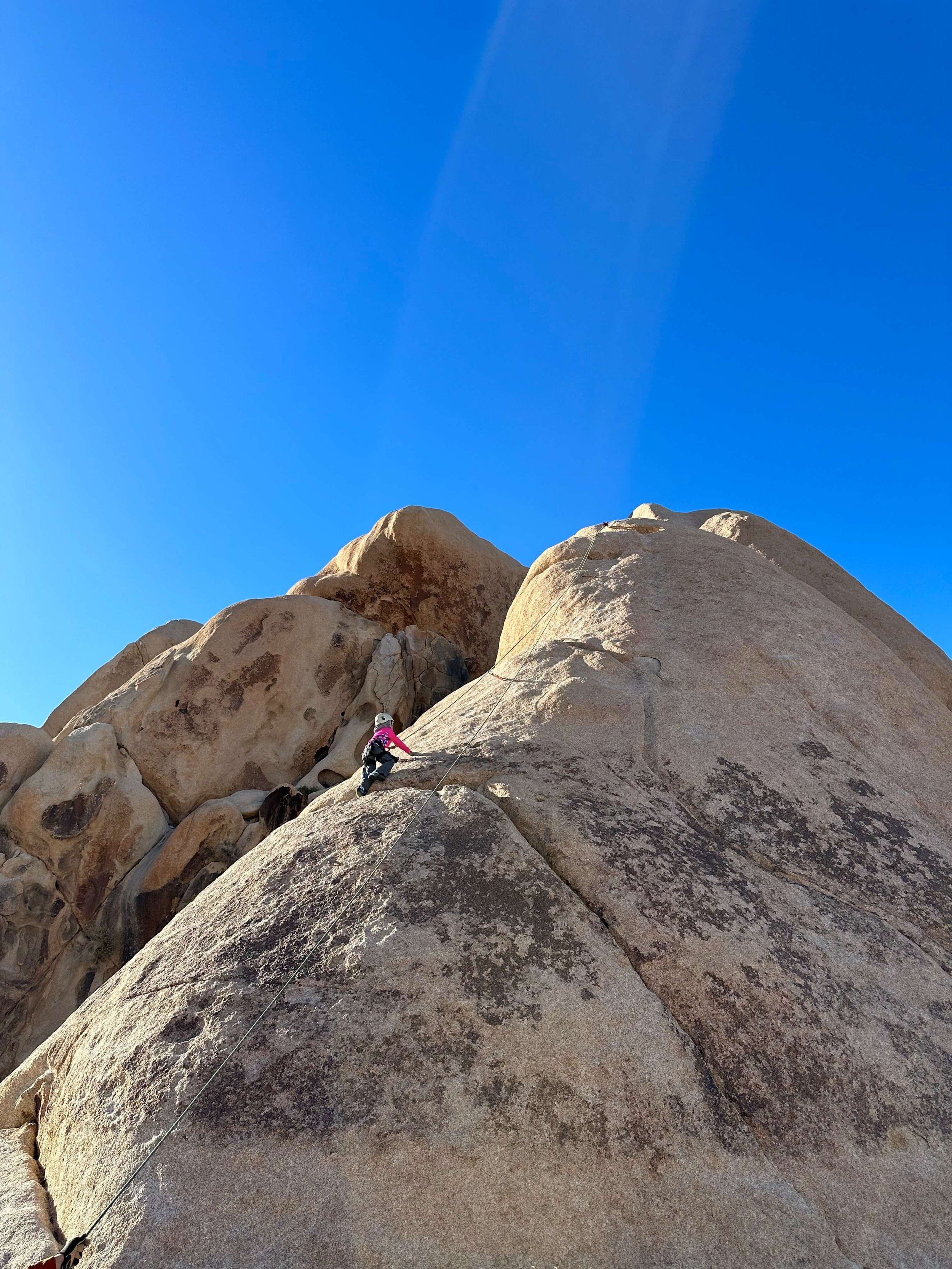 Child in pink jacket and white helmet rock climbing on large granite boulder under clear blue sky.