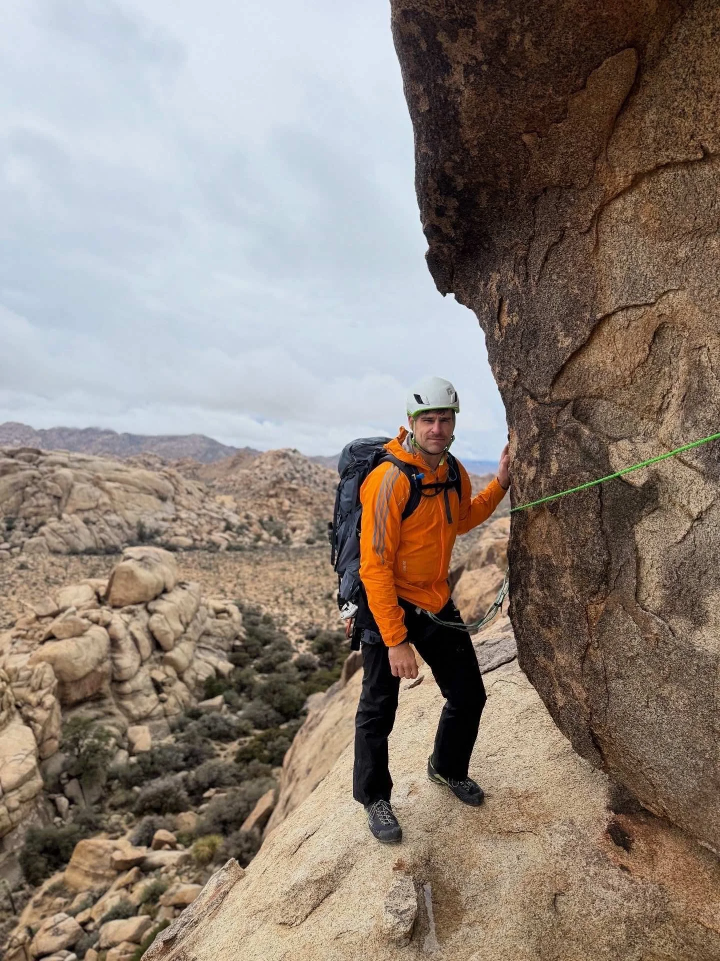 A man wearing an orange jacket, black pants, climbing shoes, a helmet, and a harness is rock climbing on a large boulder in a desert landscape with rocky hills and bushes in the background.