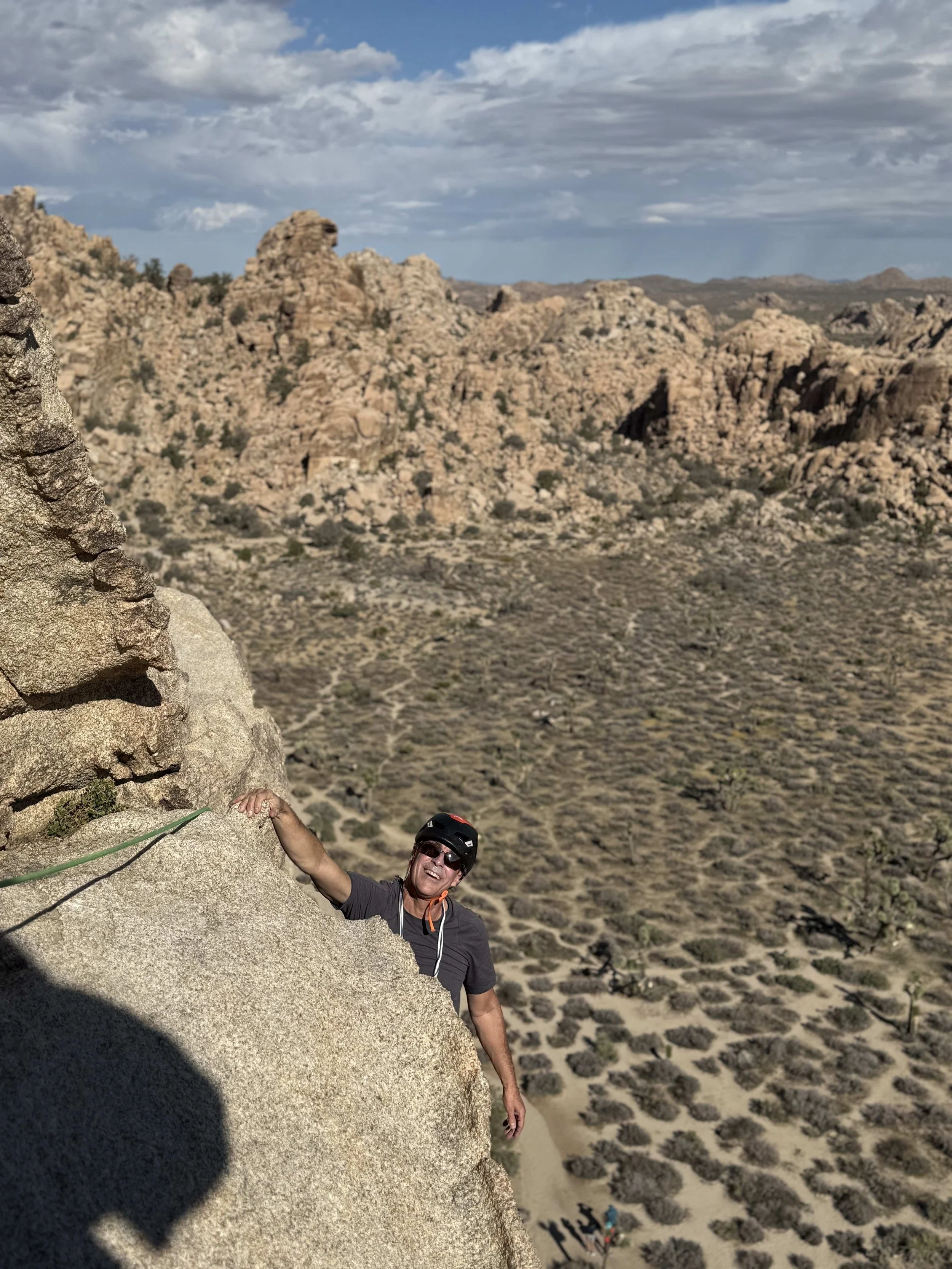 A man wearing a helmet and sunglasses climbing a steep rock wall in an arid desert landscape with rocky hills and sparse vegetation.