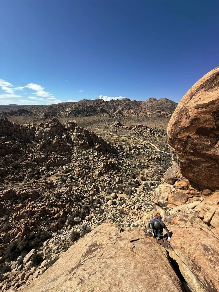 A hiker climbing a large rock formation in a desert landscape with mountains and a clear blue sky in the background.