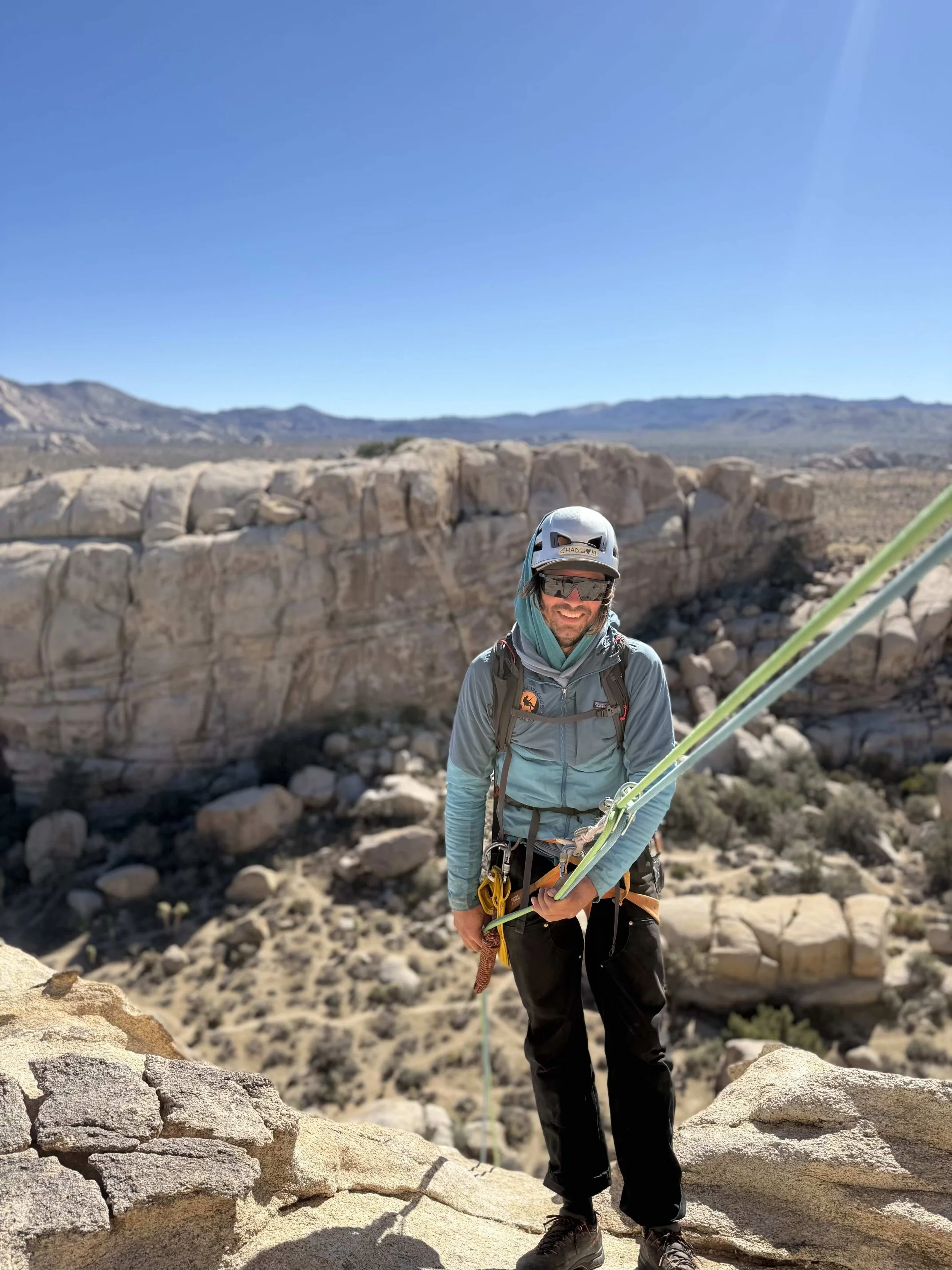 A male rock climber in a helmet and sunglasses standing on a rocky ledge in a desert landscape with large boulders and distant mountains under a clear blue sky.