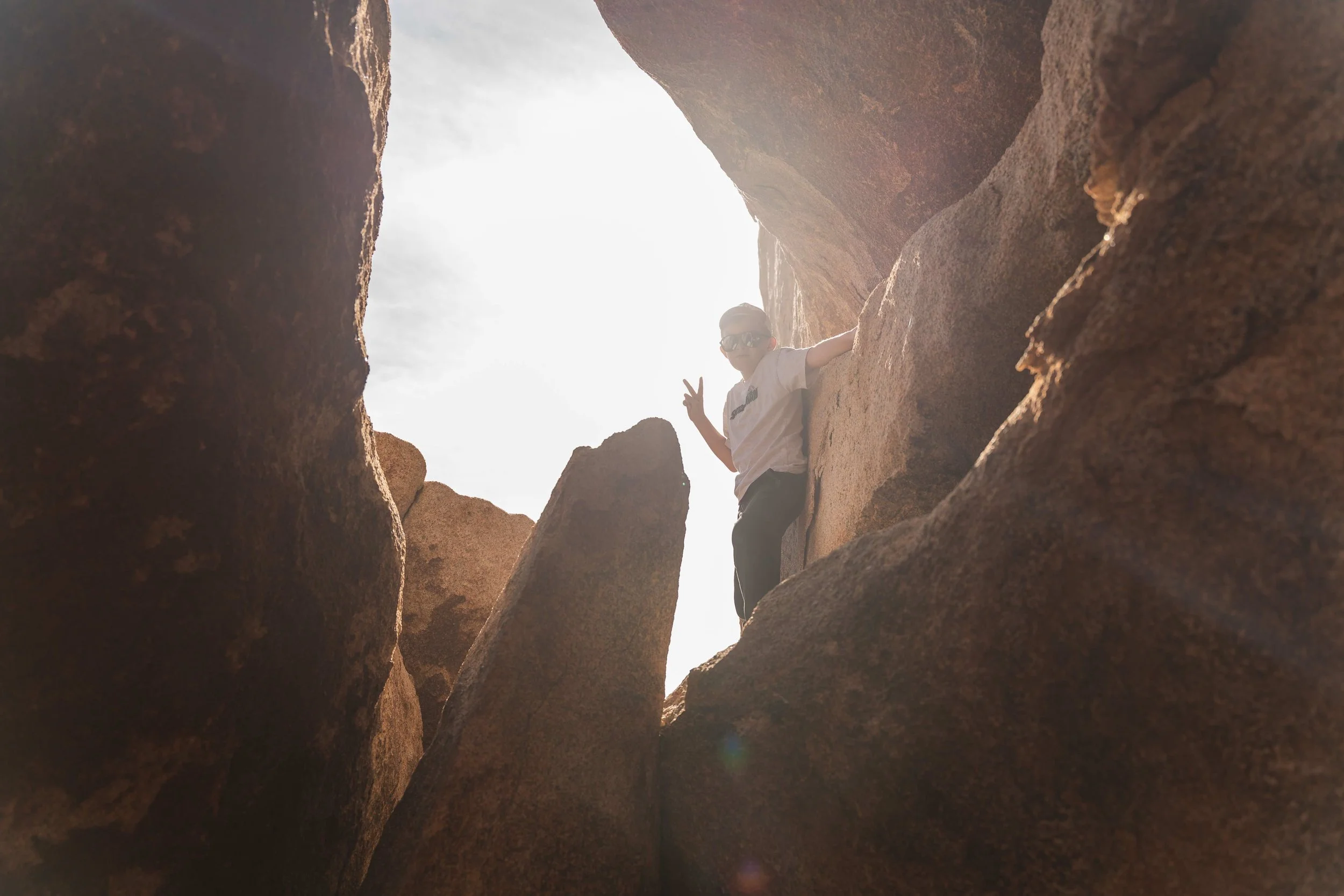 A child wearing sunglasses and a white T-shirt making a peace sign while climbing or sitting among large rocks or cliffs outdoors during daytime.