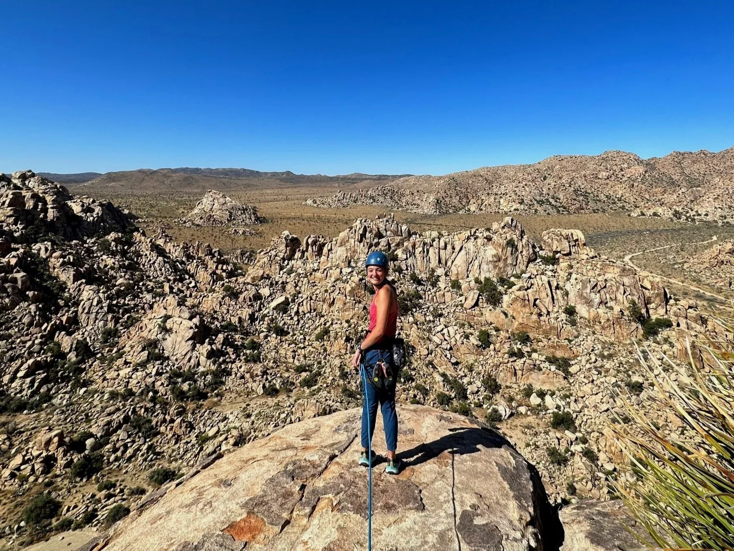A woman wearing a helmet and climbing harness stands on a large rock at a desert mountain range, smiling with rocky hills and a clear blue sky in the background.