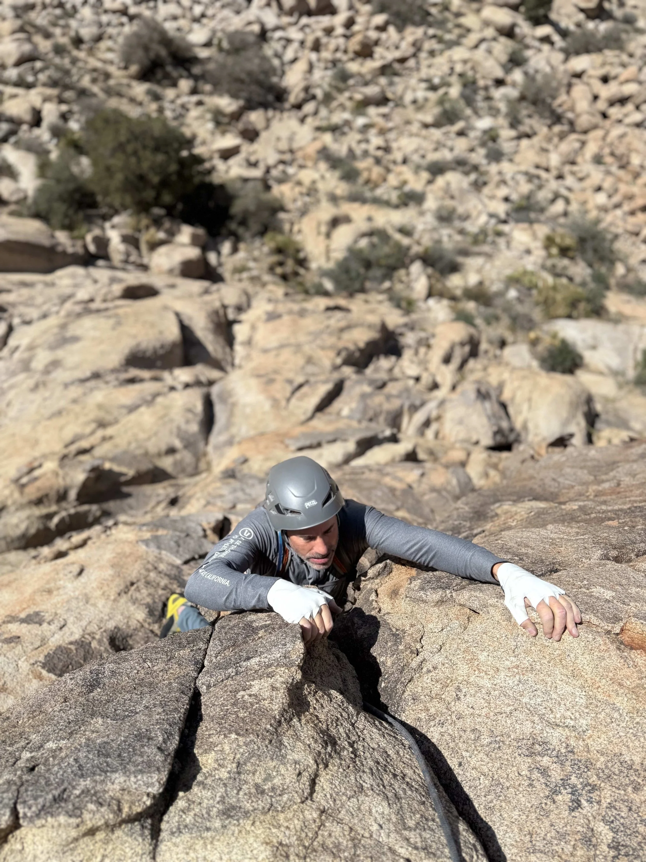 A man in a gray helmet and long sleeve shirt is climbing a steep rocky terrain, using both hands for support during a rock climb in a desert environment.