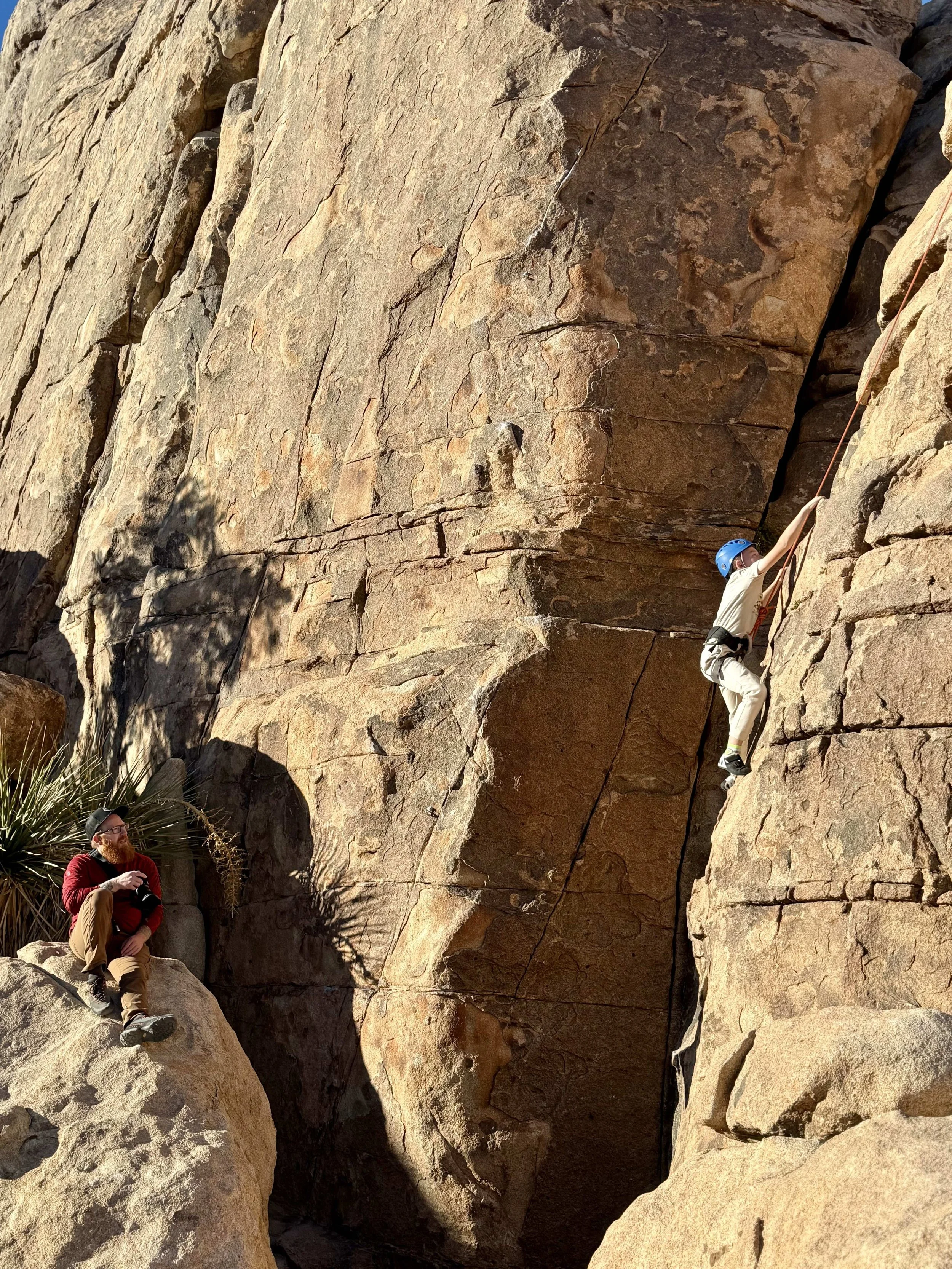 A person wearing a blue helmet and climbing gear is climbing a tall rock face, while another person with a beard, hat, and red sweater is sitting on a large rock watching and taking notes.
