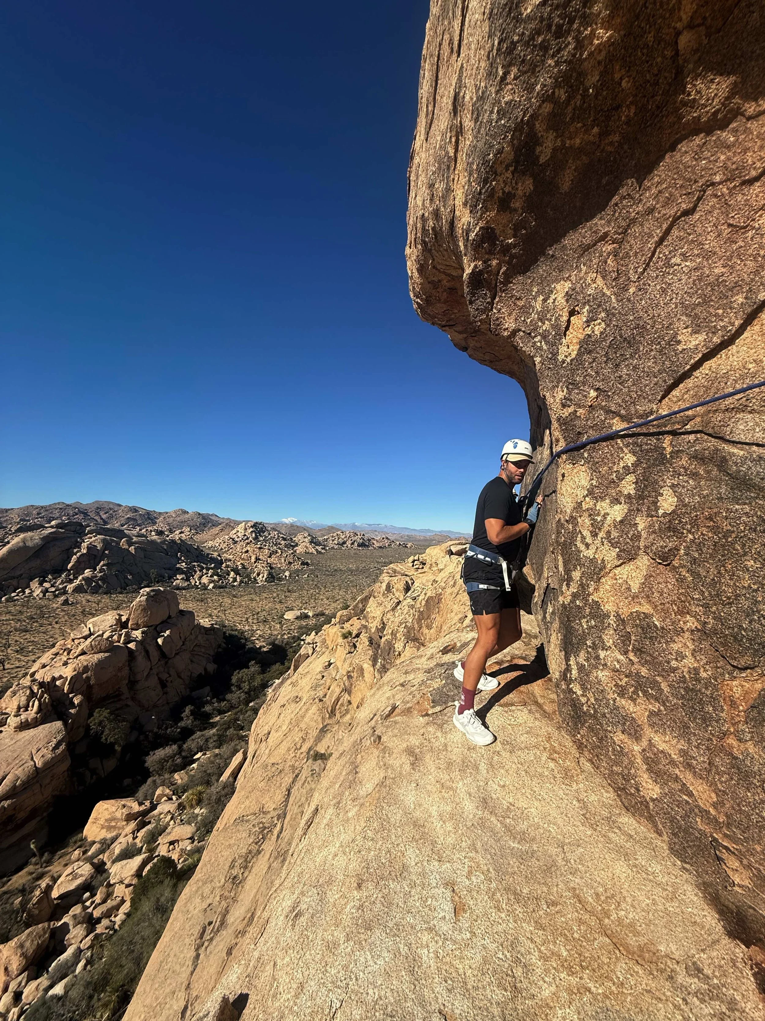 A person wearing a helmet and harness rock climbing on a large sandstone rock formation in a desert landscape.