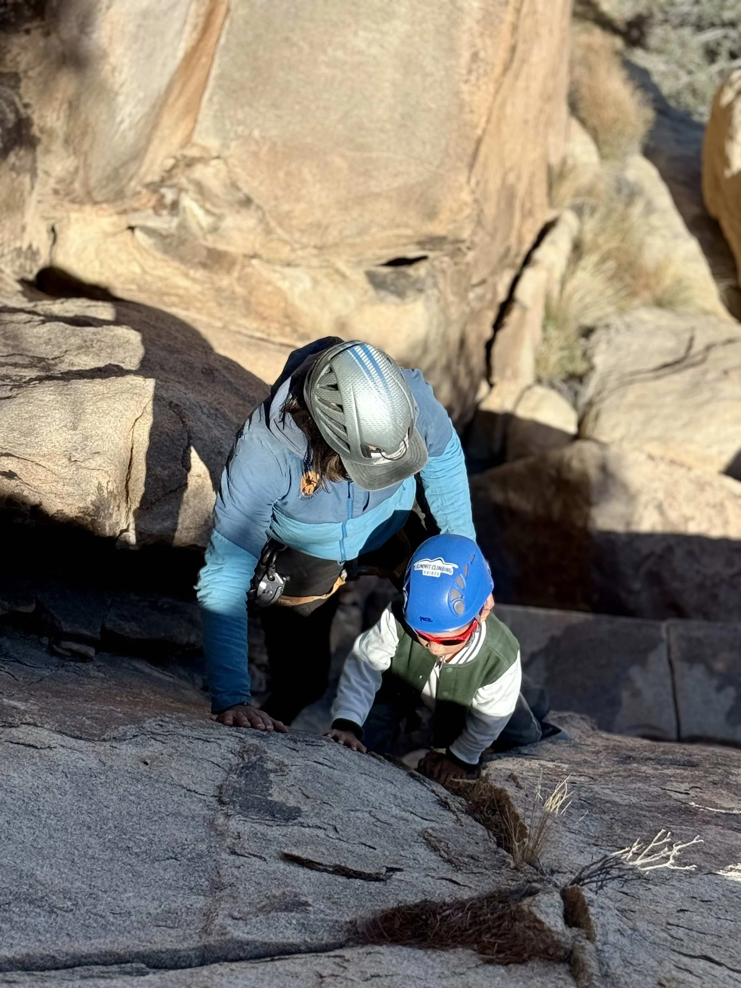 An adult and a child rock climbing together on a steep rocky surface, both wearing helmets and outdoor climbing gear.