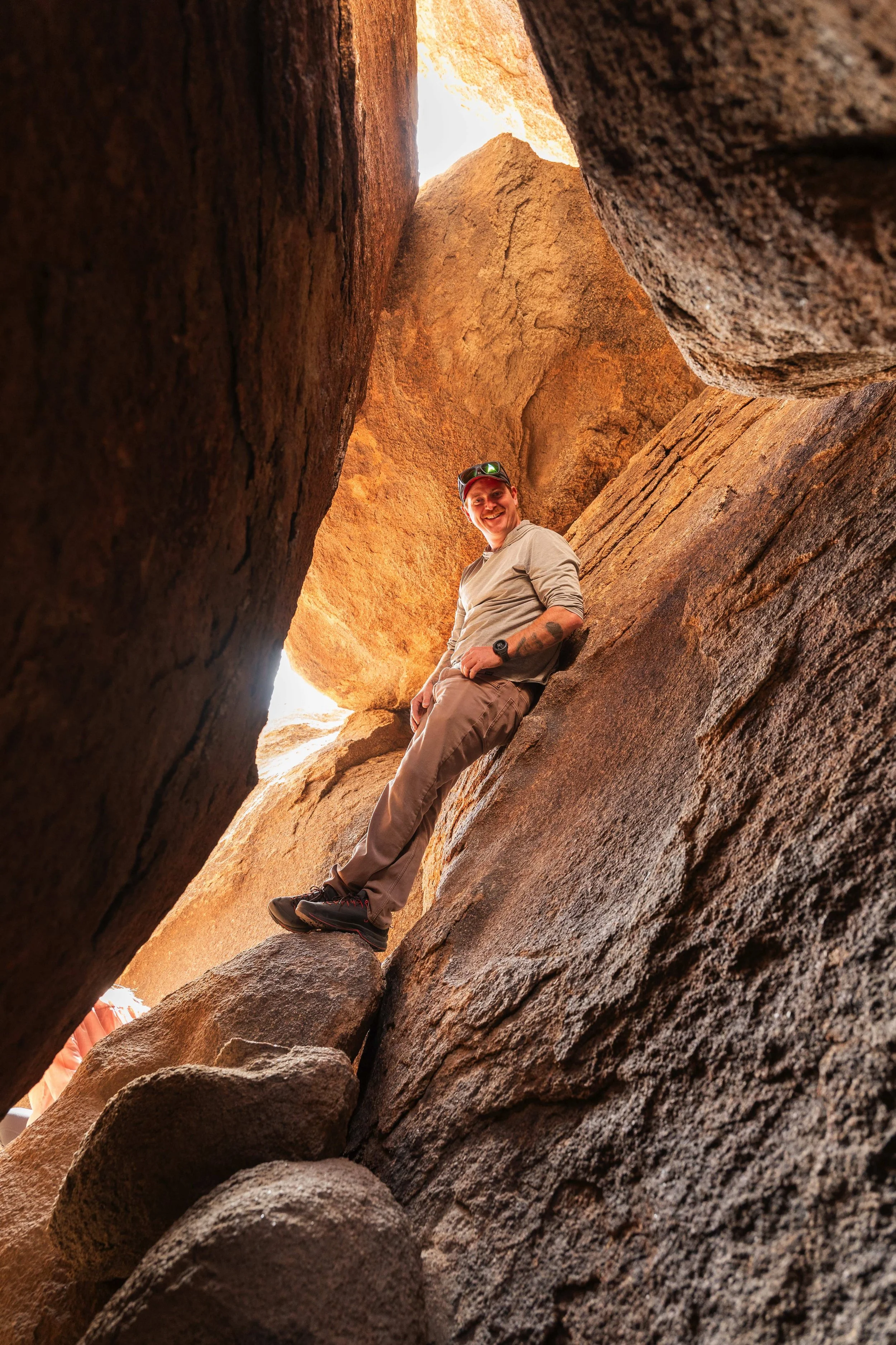 A man smiling while sitting on a large rock between tall, reddish-brown boulders.