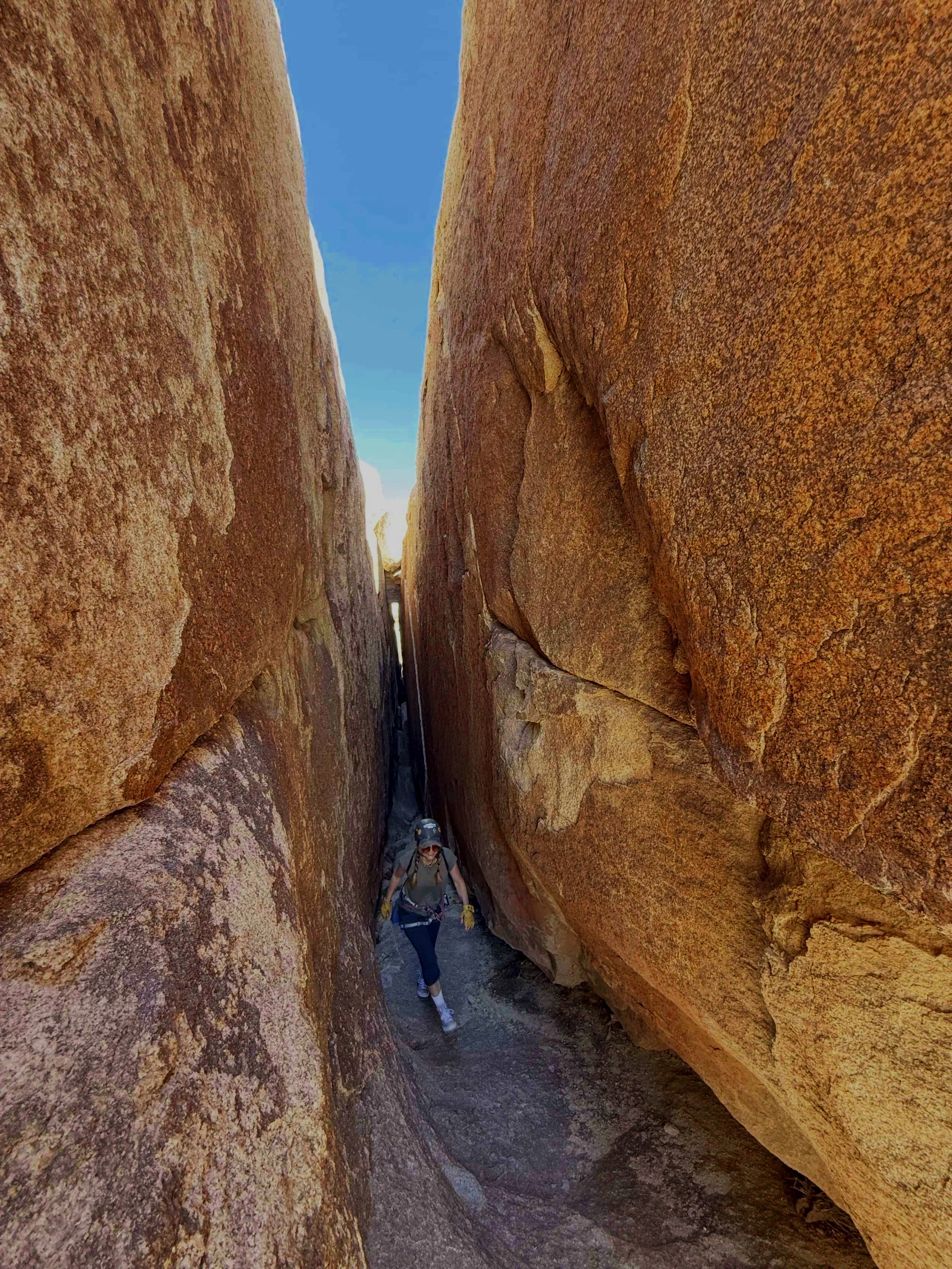 A person with a backpack and hat hiking through a narrow slot canyon with large reddish-brown rock walls.