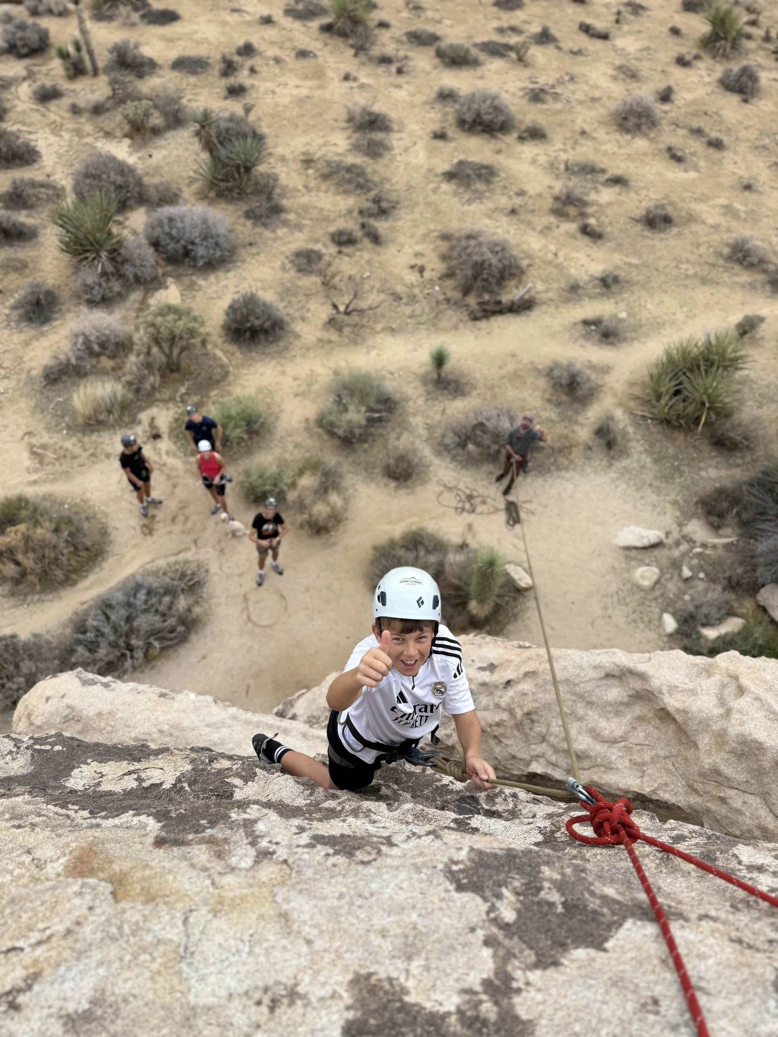 Child rock climbing on a rock wall wearing a helmet, with a smiling face giving a thumbs-up. Ropes and other people are visible below in a desert landscape.