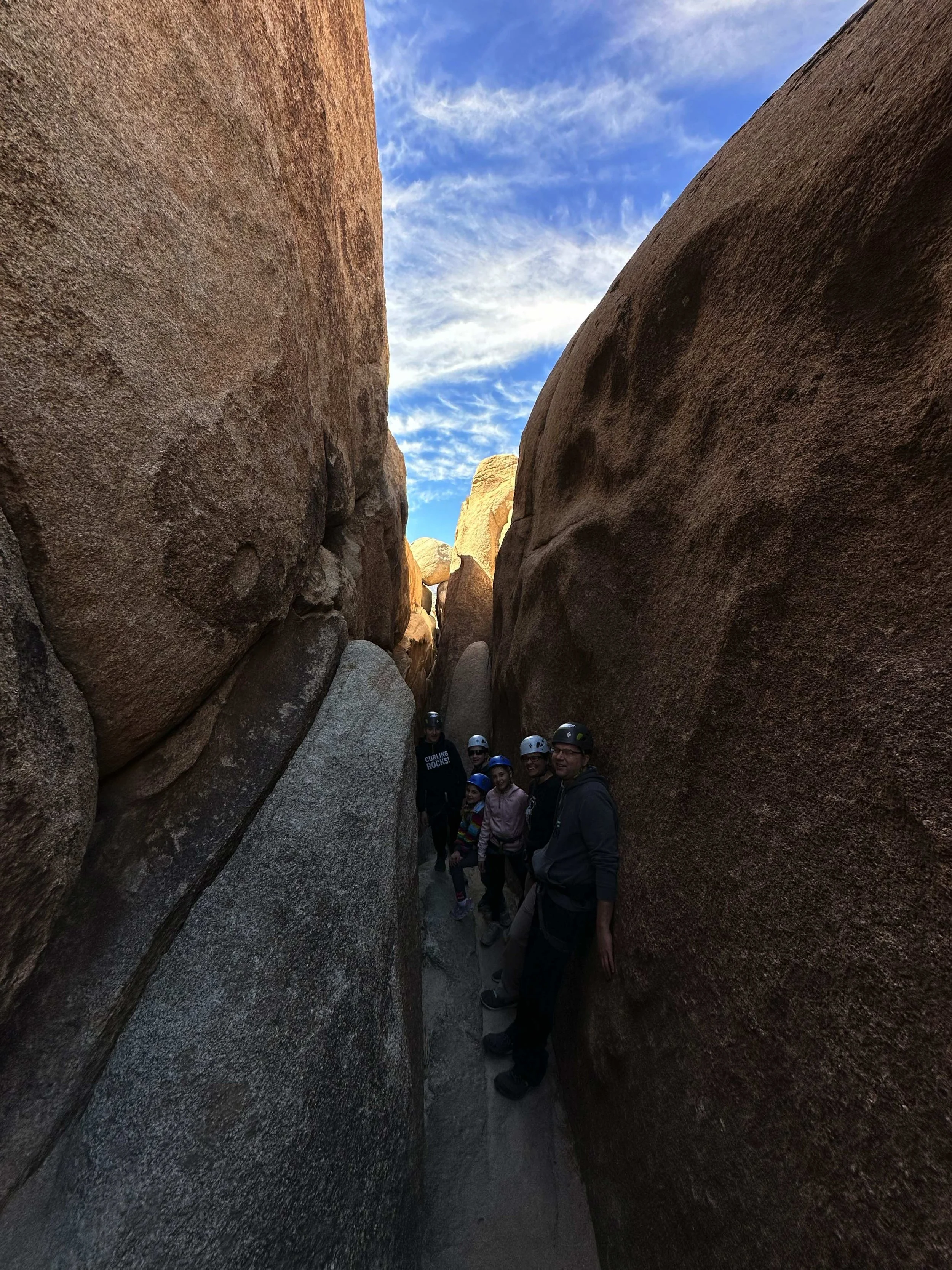 A group of people wearing helmets standing in a narrow slot canyon with large rock walls on either side and a partly cloudy sky above.