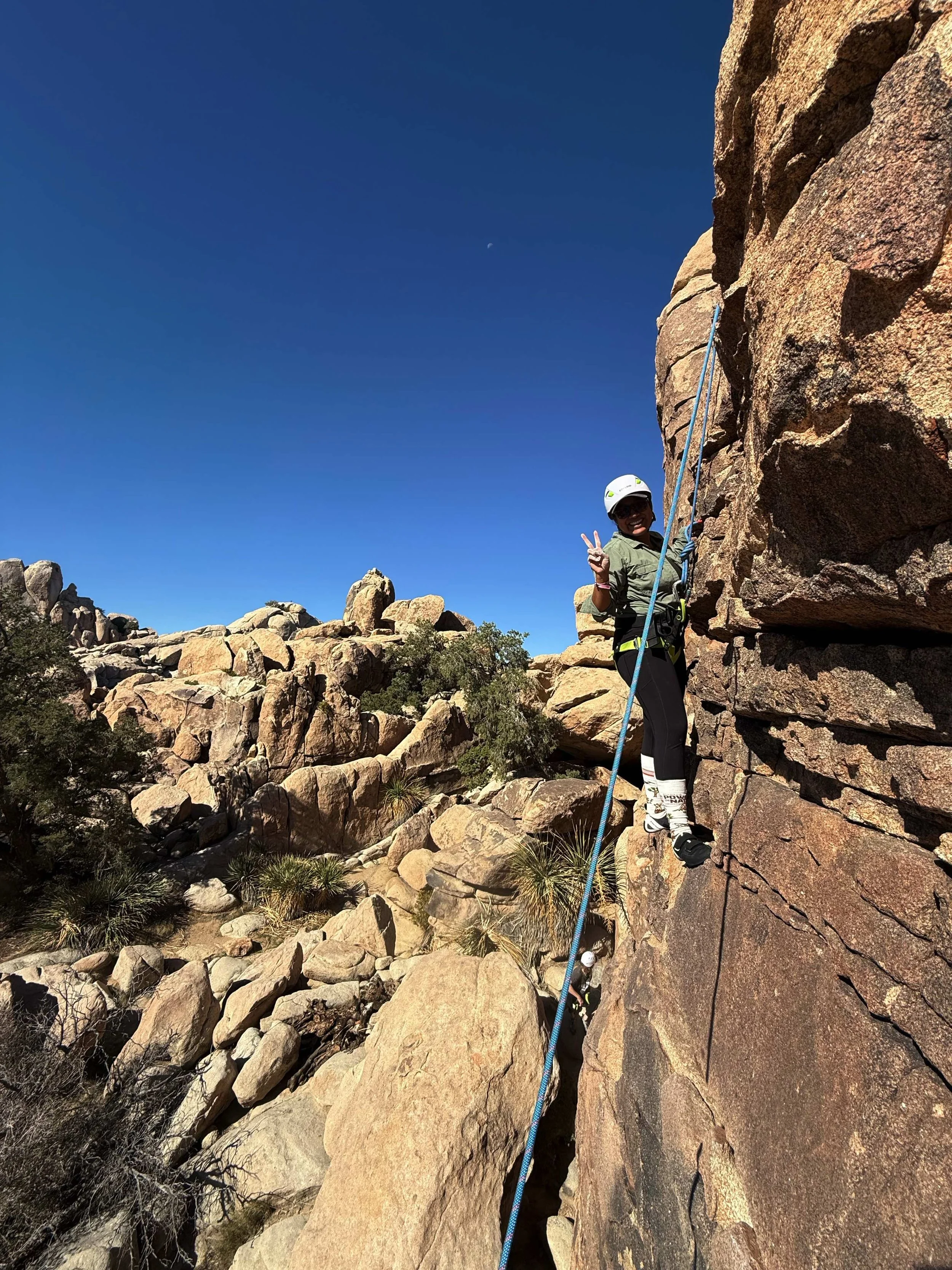 Person rock climbing on a steep rock face, wearing a helmet and harness, smiling and making a peace sign with their hand in a desert landscape with large boulders and sparse vegetation under a clear blue sky.