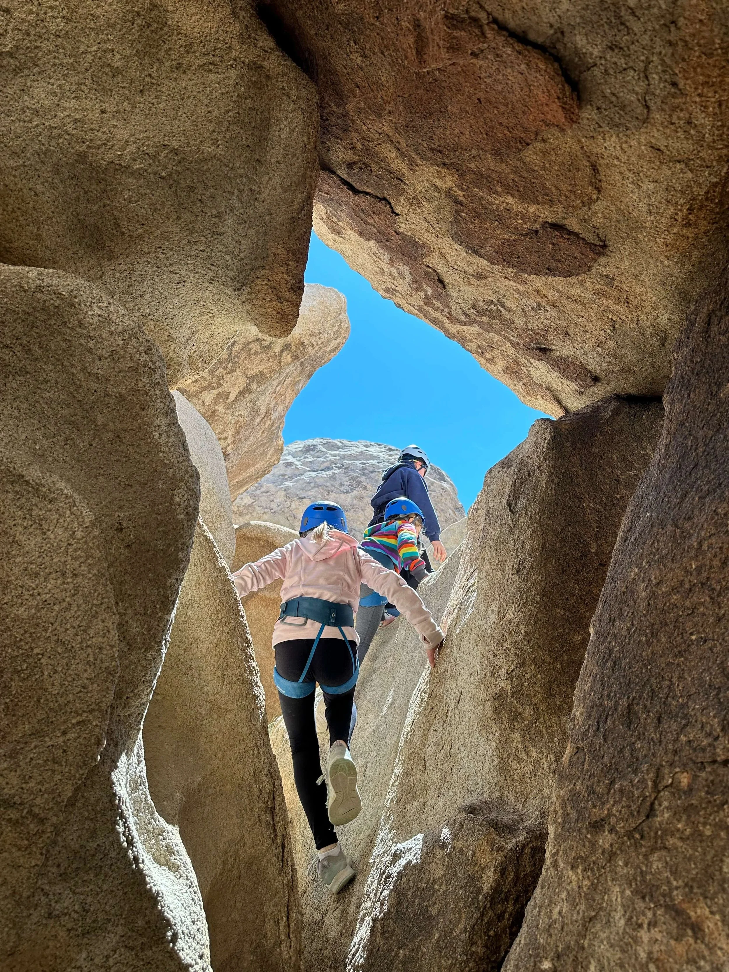 People climbing through a narrow rock canyon, wearing helmets and harnesses, with a blue sky overhead.