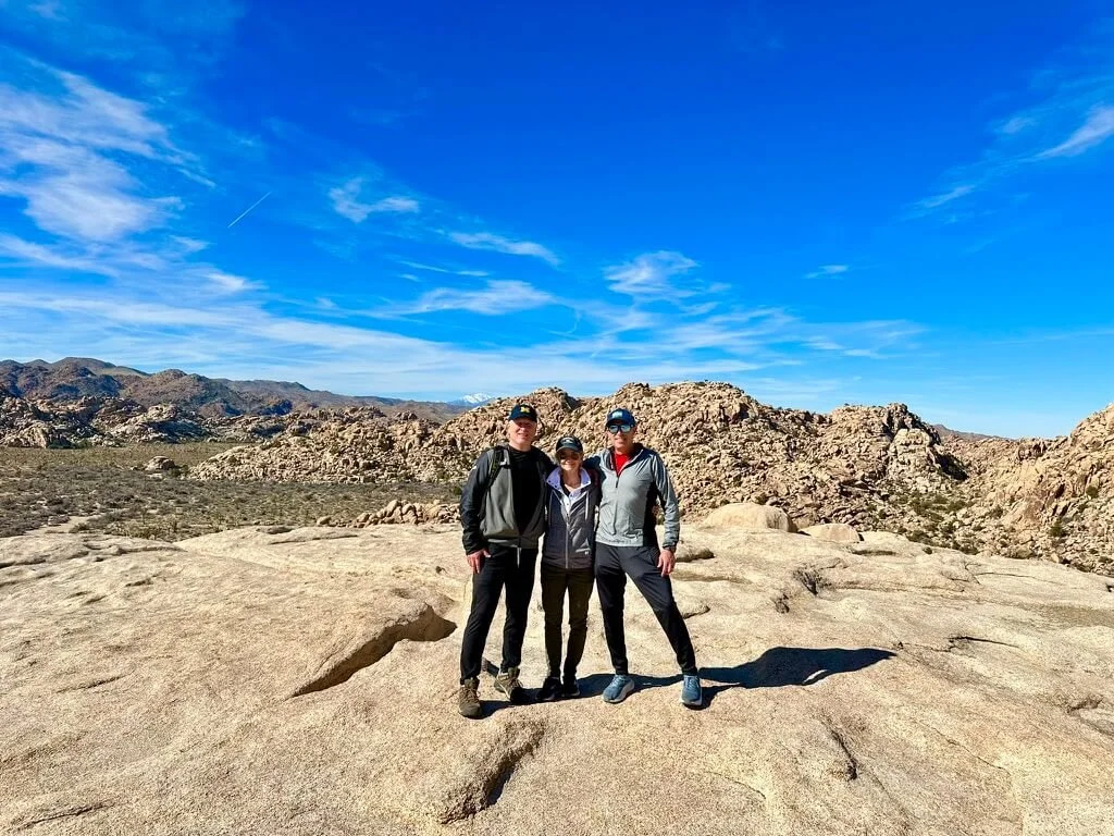 Three people standing on rocky terrain in a desert landscape with mountains and a clear blue sky in the background.