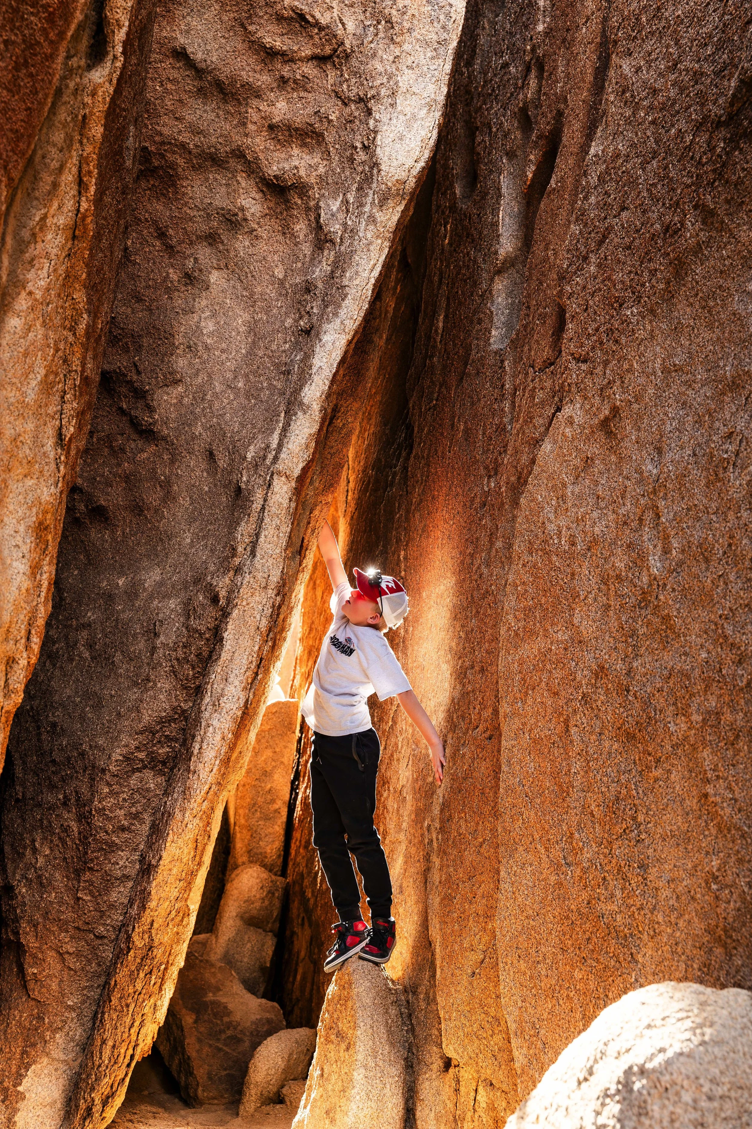 A young boy wearing a white T-shirt, black pants, and red shoes is climbing among large rugged boulders in a desert or canyon setting, with sunlight illuminating the scene.