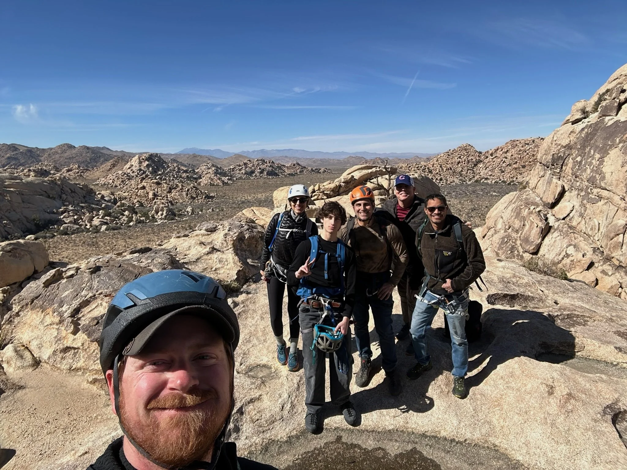 Group of six people, some wearing helmets, hiking in a desert landscape with rocky terrain and mountains in the background under a clear blue sky.